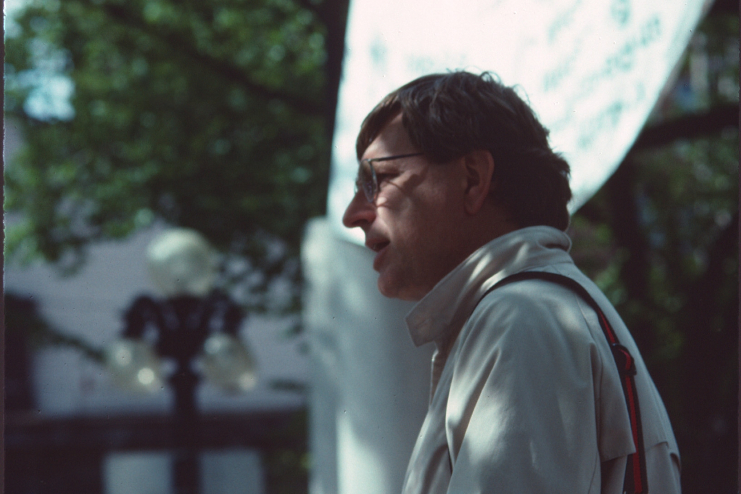 A man in a light jacket stands outdoors under trees, with a streetlamp and blurred banner in the background.