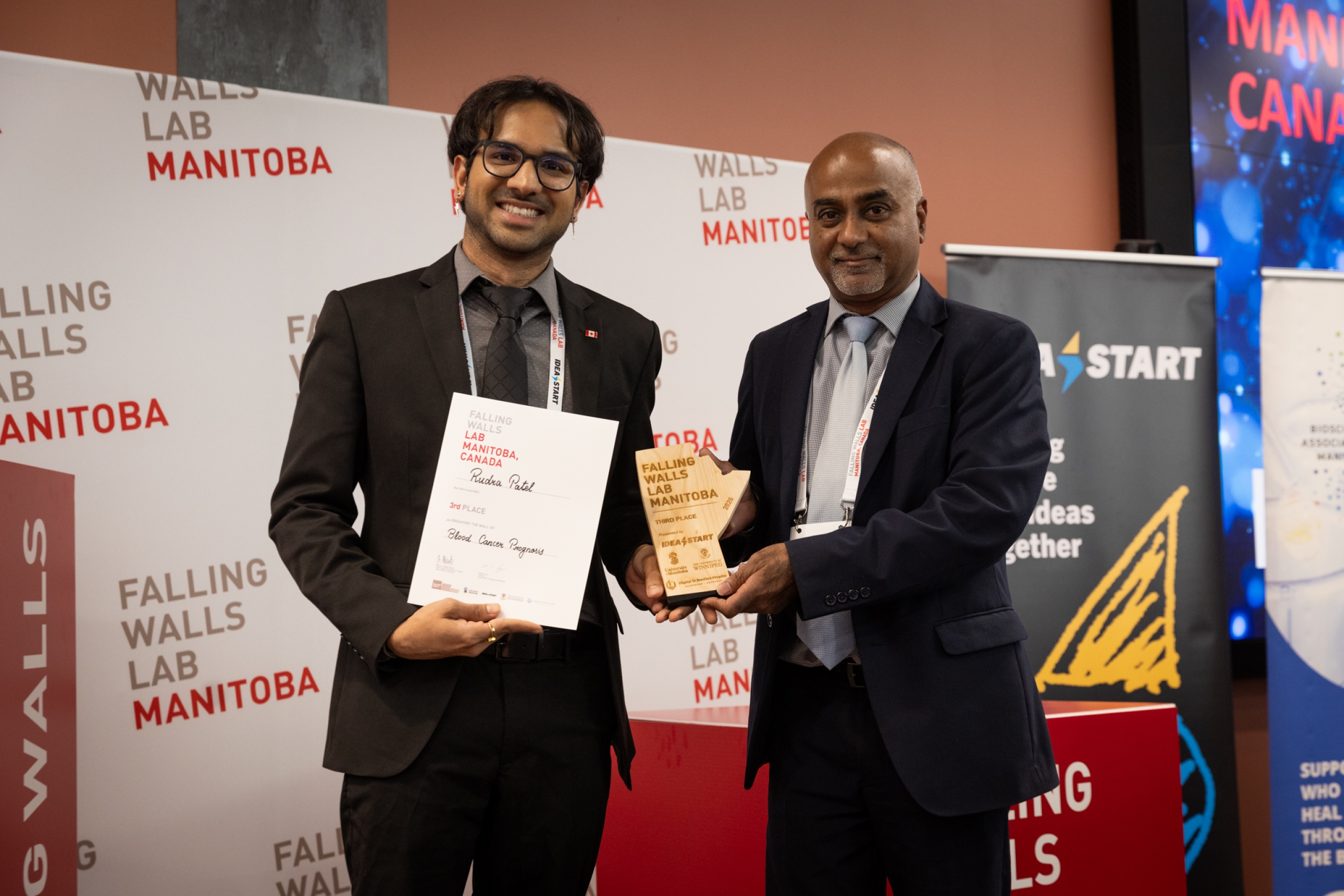 Third place winner stands with an award presenter, holding a certificate and trophy at Falling Walls Lab Manitoba 2026.