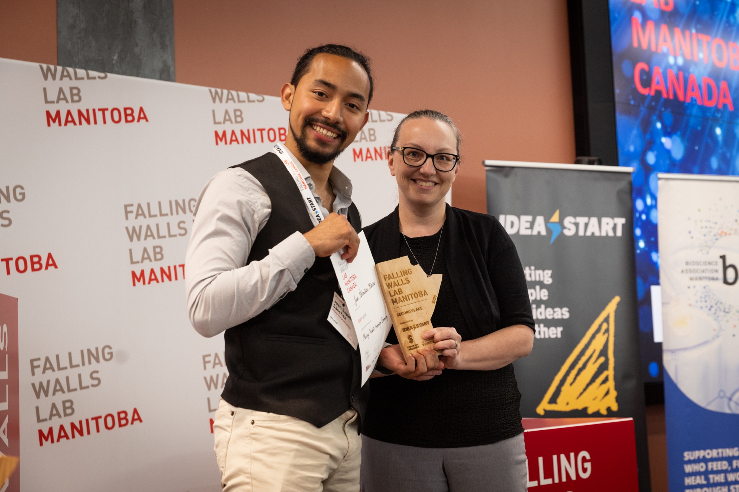 Second place winner stands with an award presenter, holding a certificate and trophy at Falling Walls Lab Manitoba 2026.