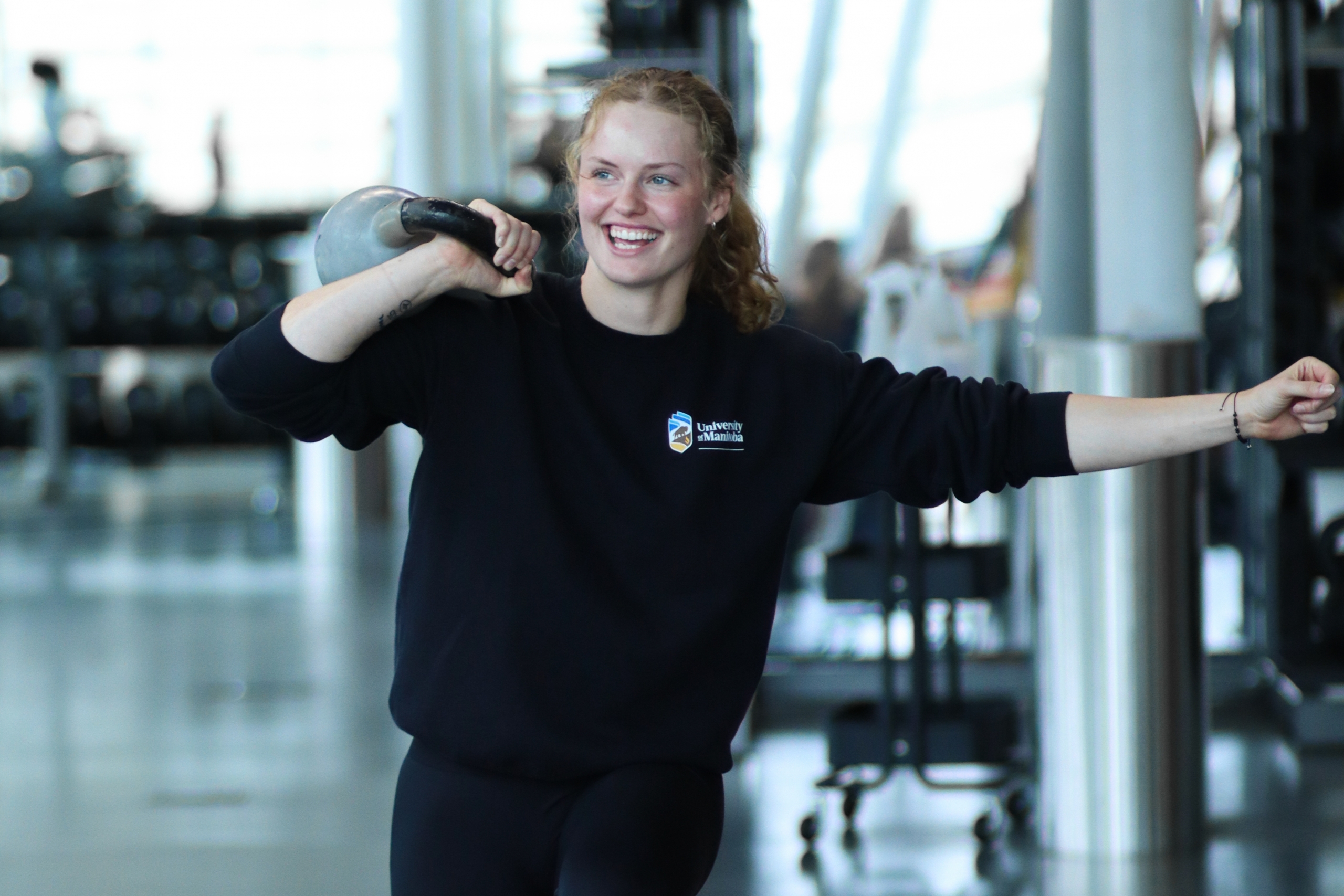 A young woman lifts a kettle bell over her right shoulder in the gym.