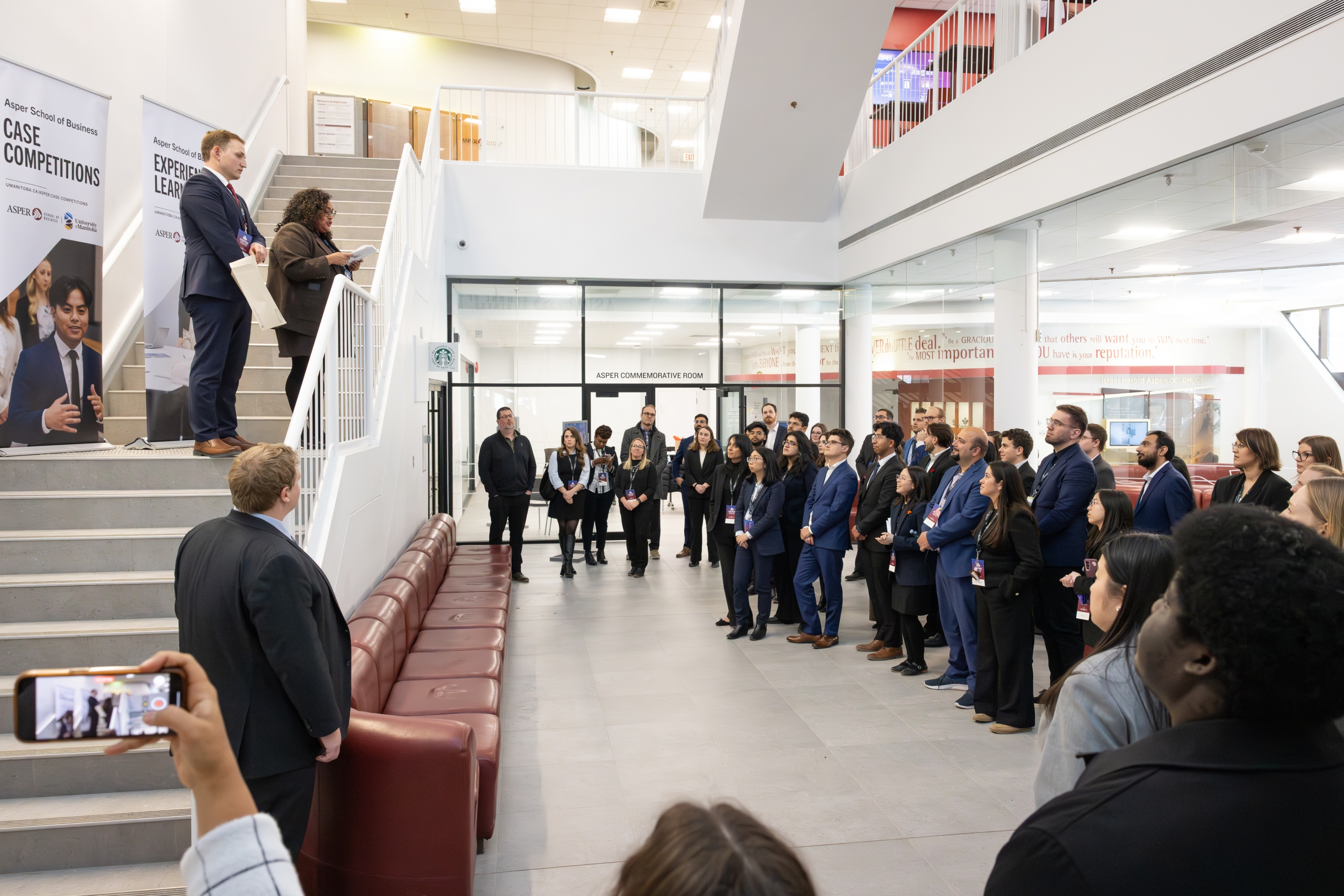 Students gather in a lobby while a speaker speaks from a staircase above