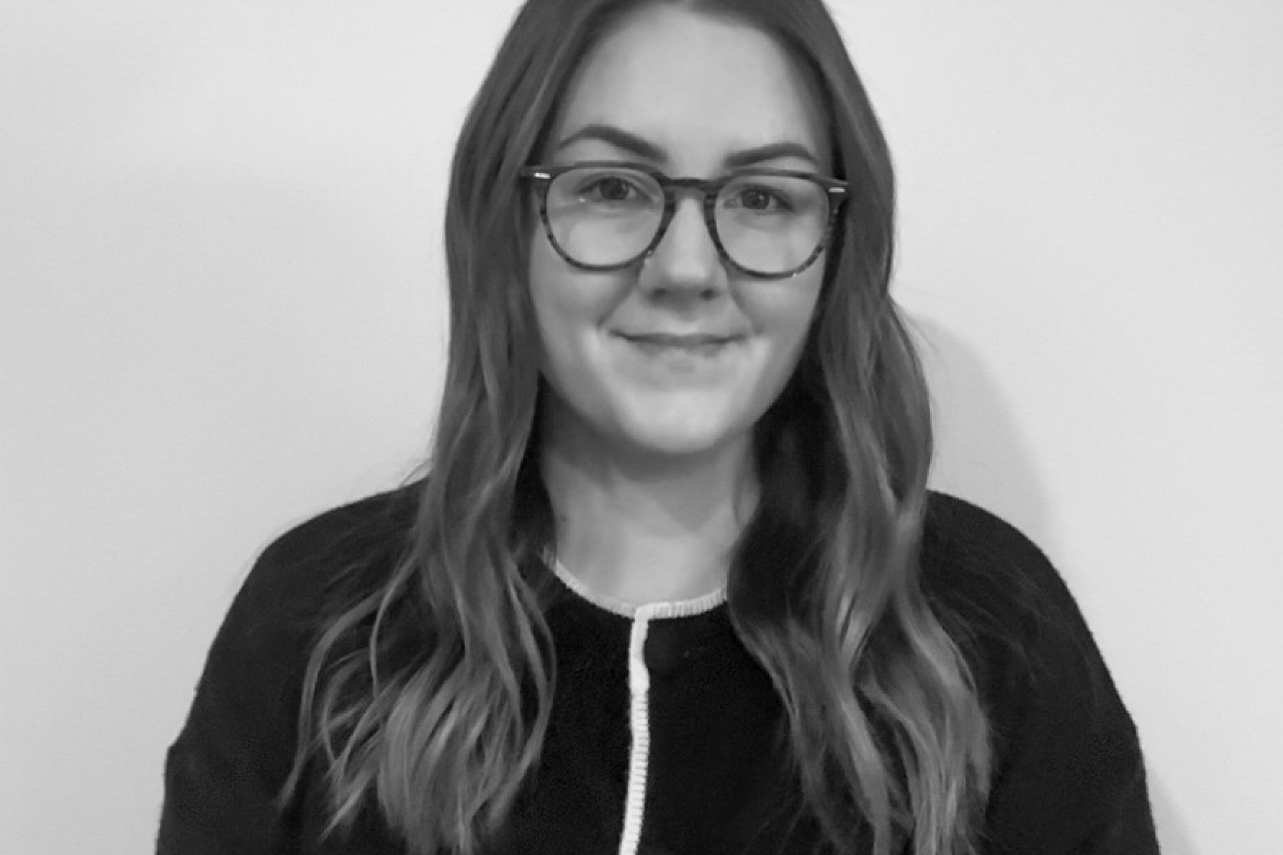 A woman with long hair and glasses smiles gently, wearing a dark cardigan with white trim.