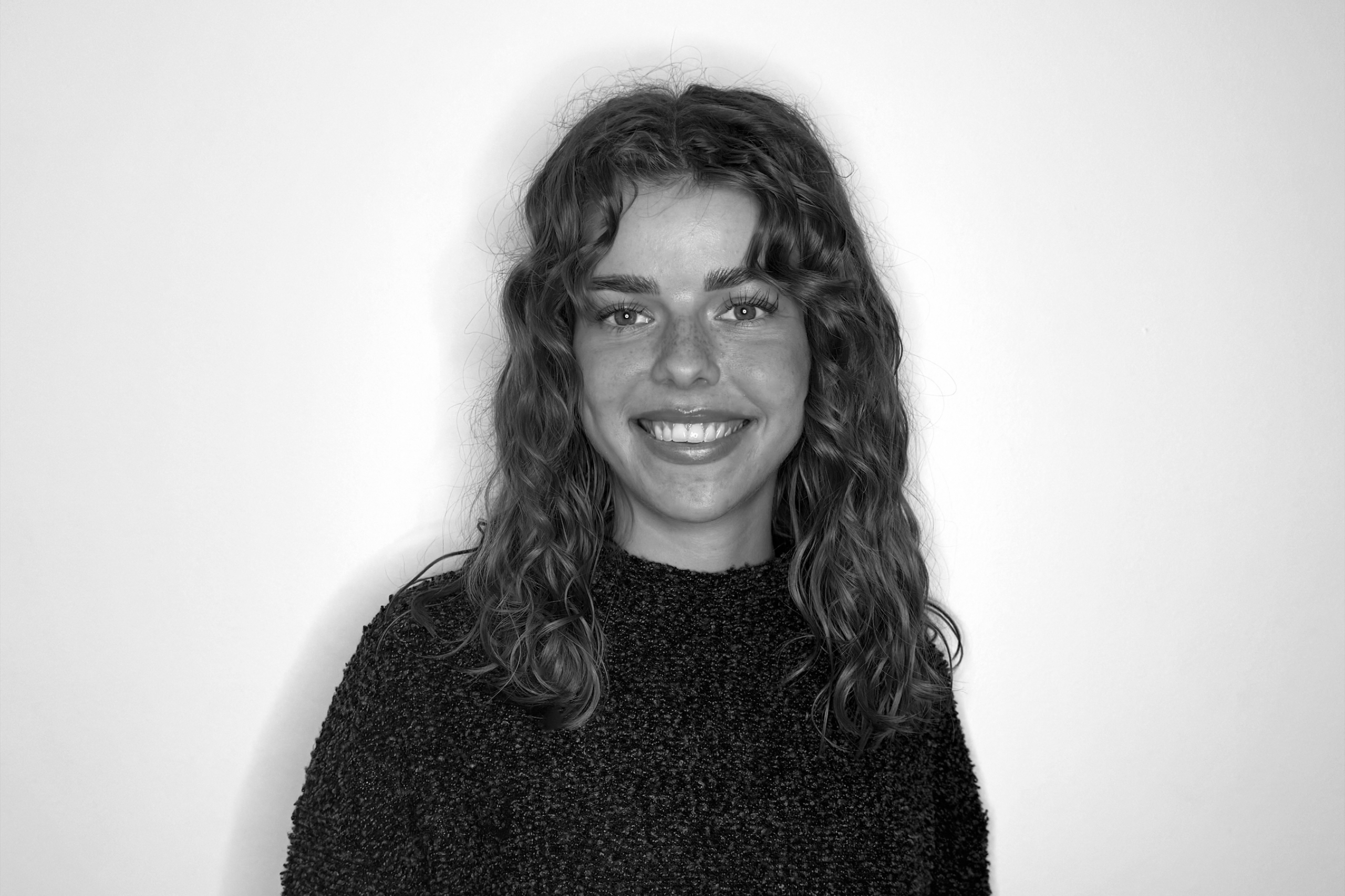 A smiling person with wavy hair wearing a dark textured sweater is centered against a plain backdrop. 