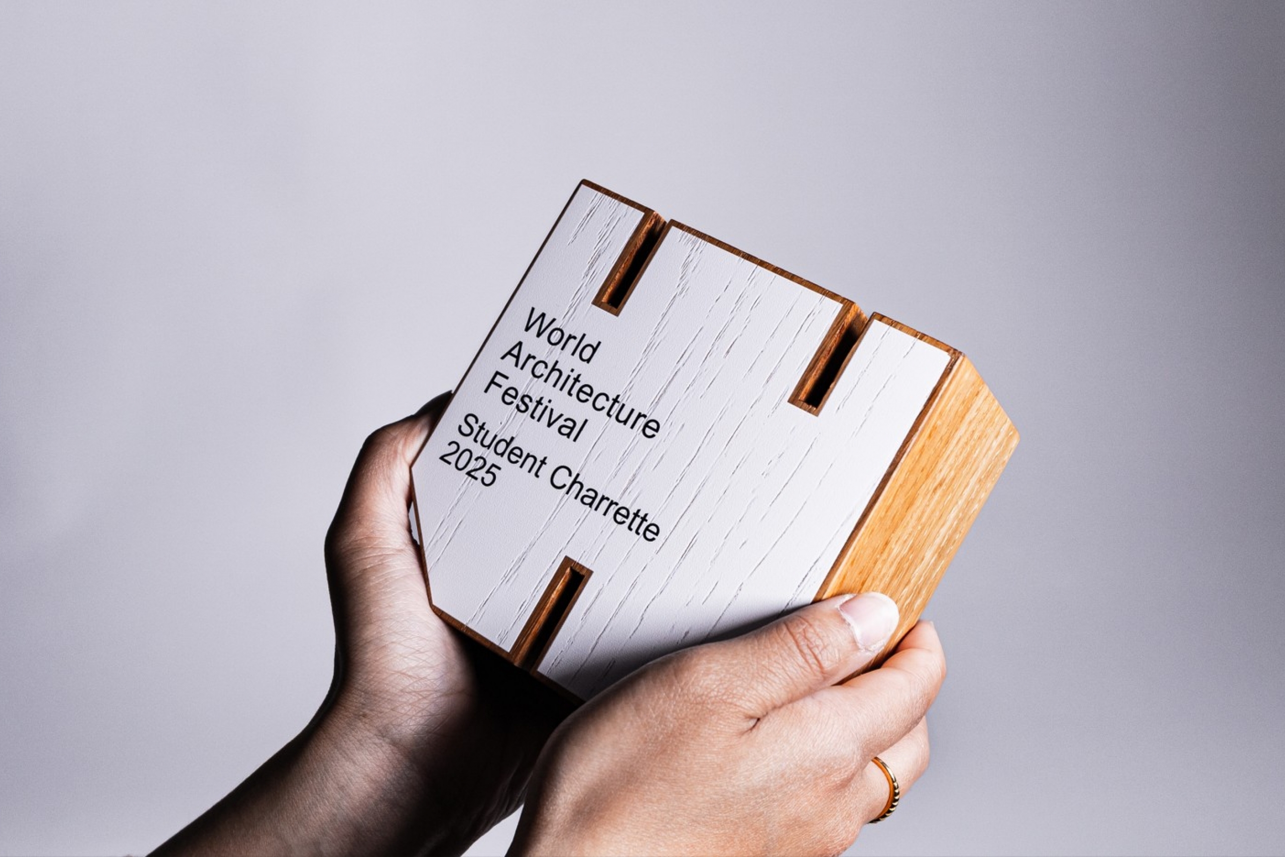 Two hands hold a white and wooden textured award inscribed with "World Architecture Festival Student Charrette 2025."