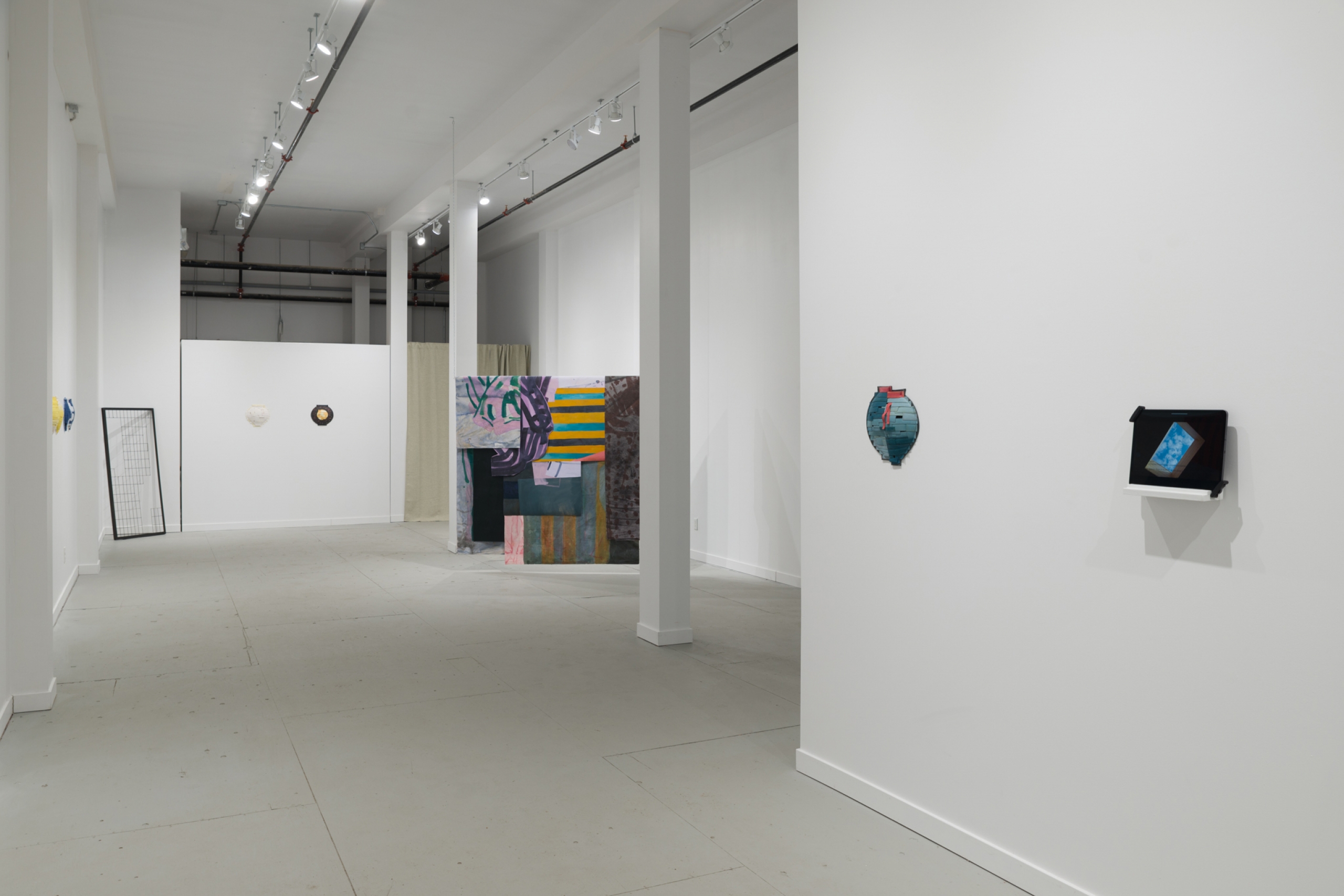 Installation view with mixed-media wall works and a colourful drapped fabric sculptural form hanging from a wooden dowel in an open gallery.