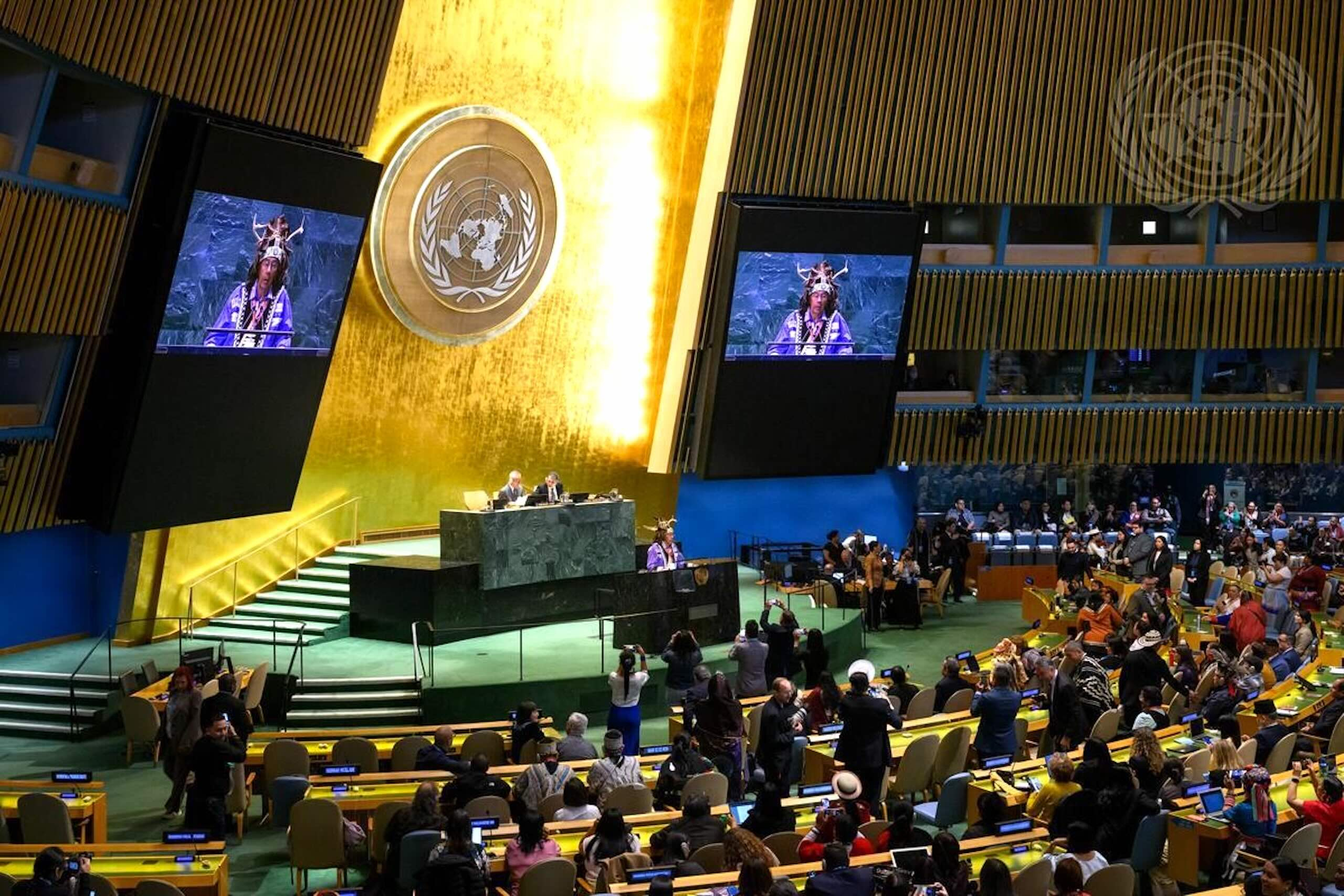 Large screens at front of room full of delegates, United Nations in New York.