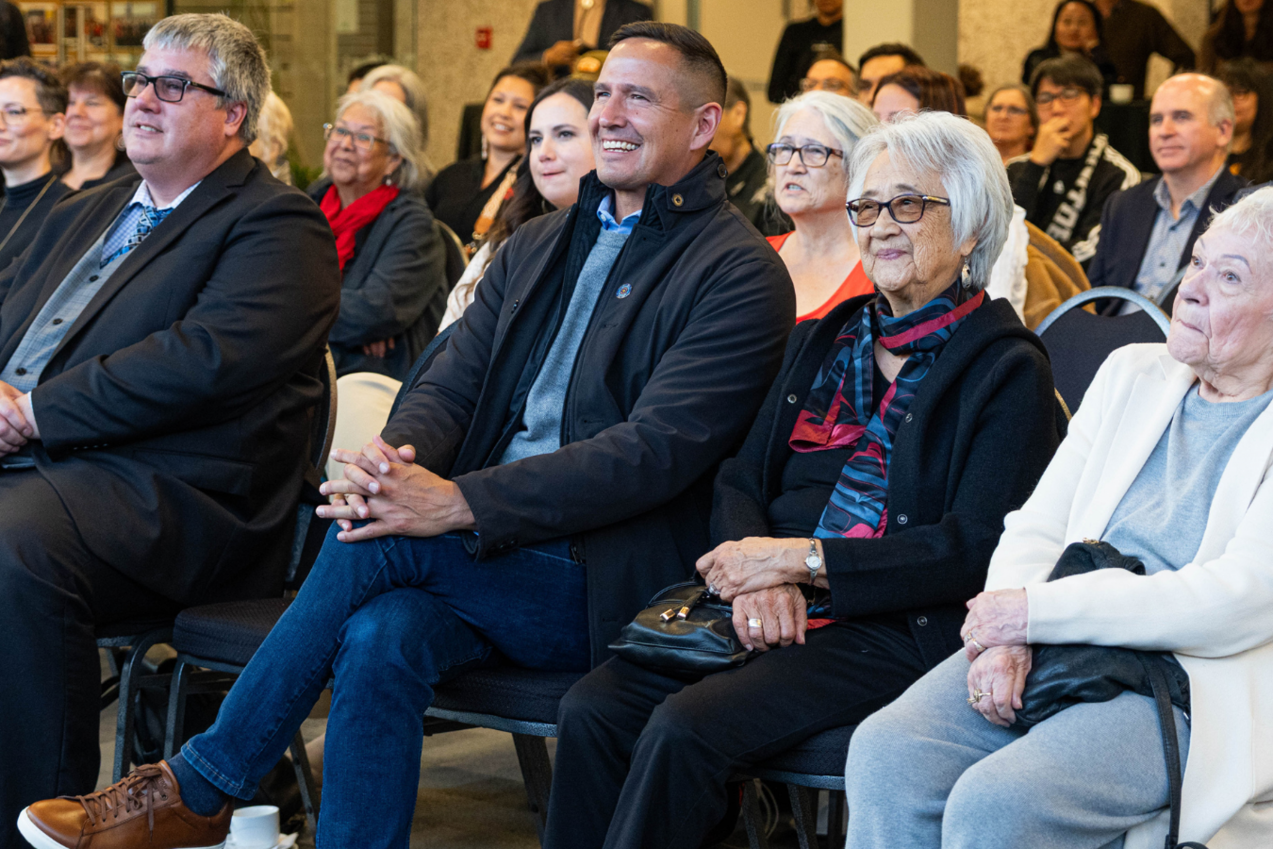 A group of attendees sitting in rows at an event, smiling and watching a presentation.