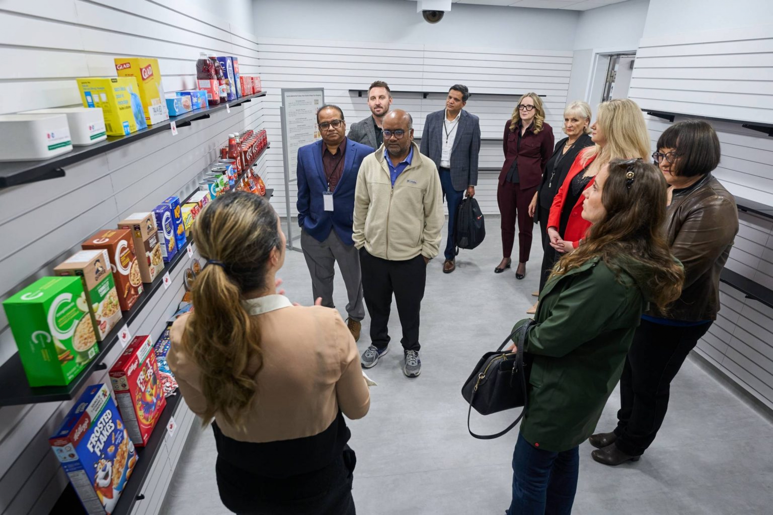A group of scholars stand in a room where each wall is made up of shelves. One shelf is lined with consumer products such as cereal and juice