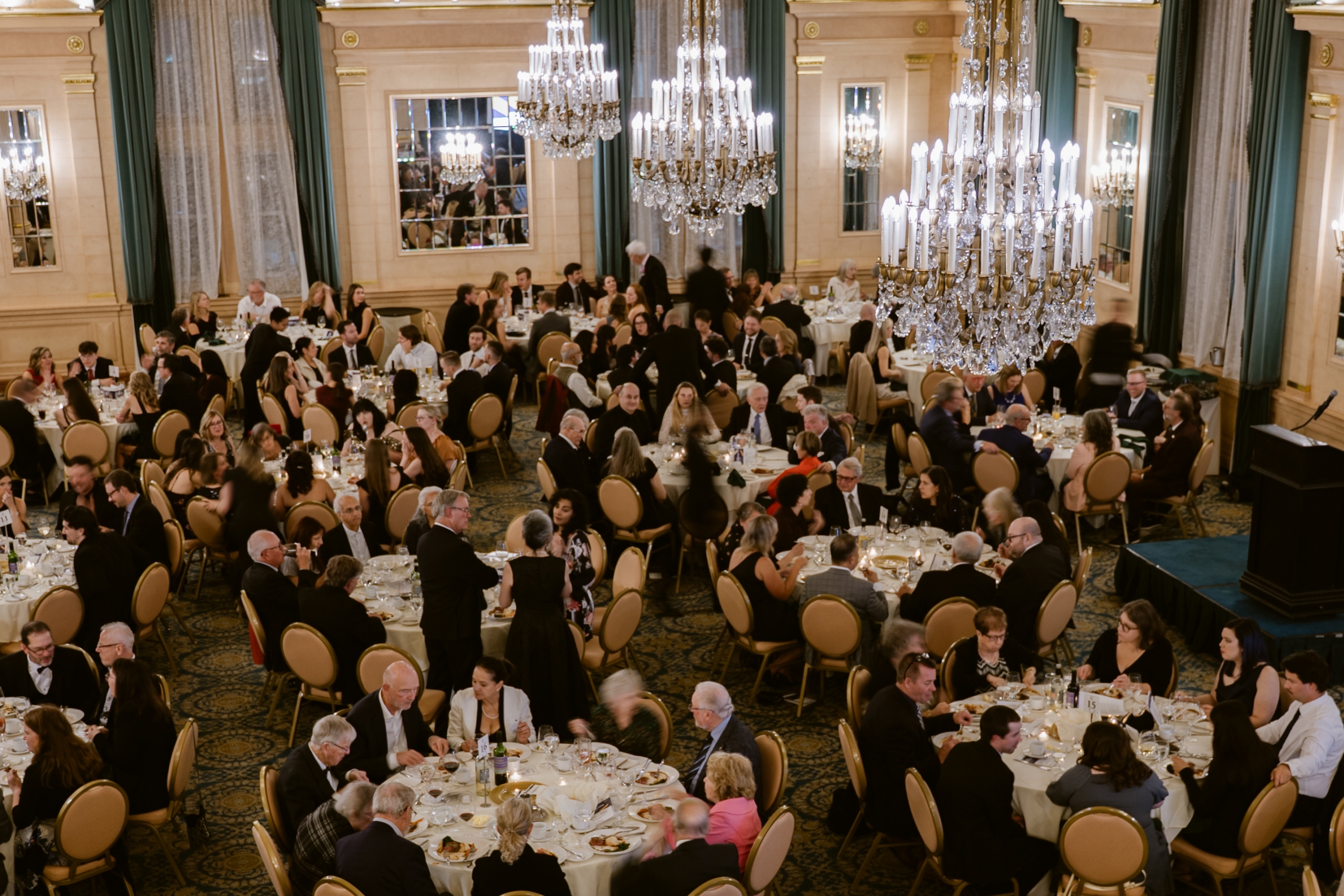 A bird's eye view of the crowd seated at round tables. Three chandeliers hang from the ceiling.