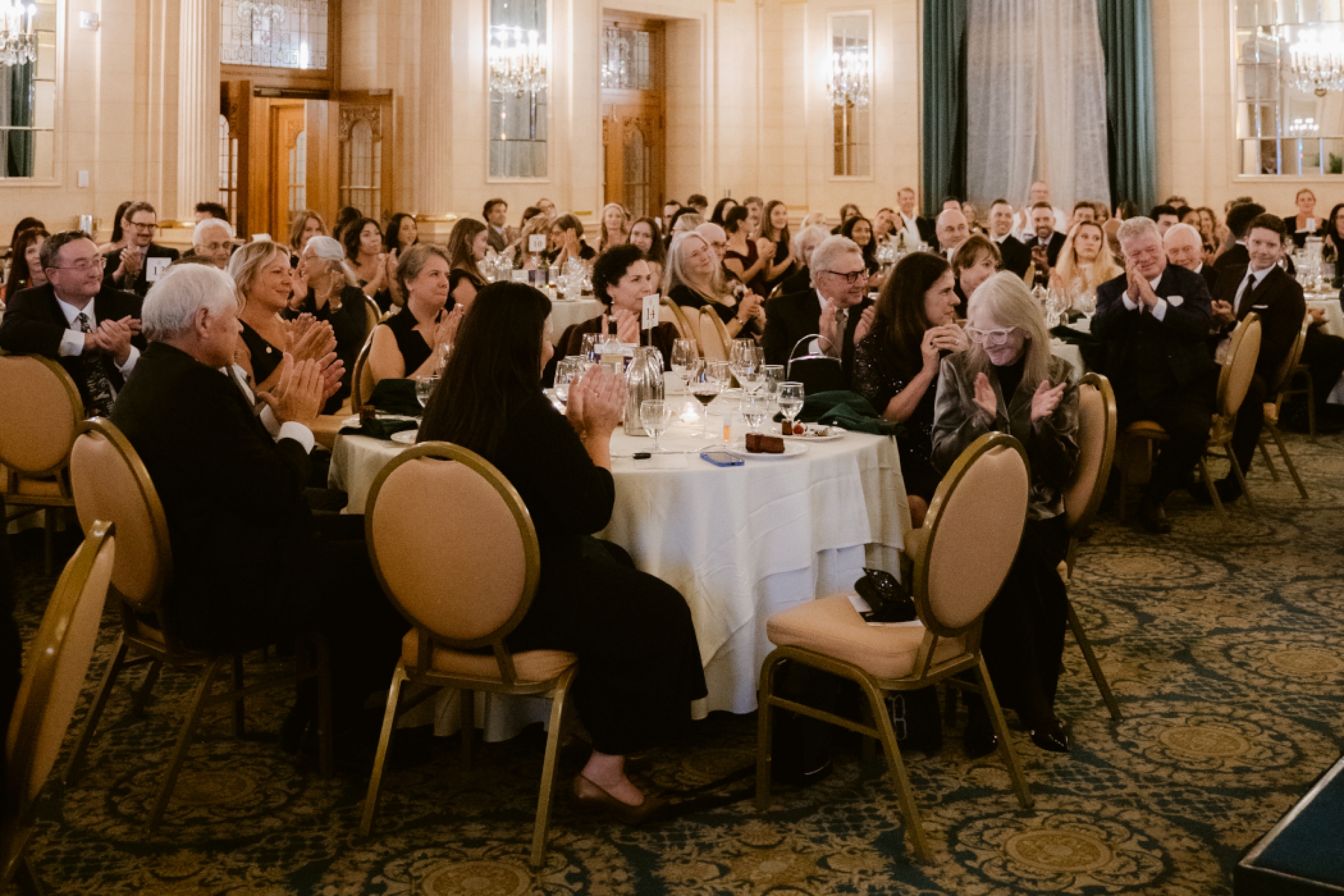 The audience, seated at round tables, claps. 