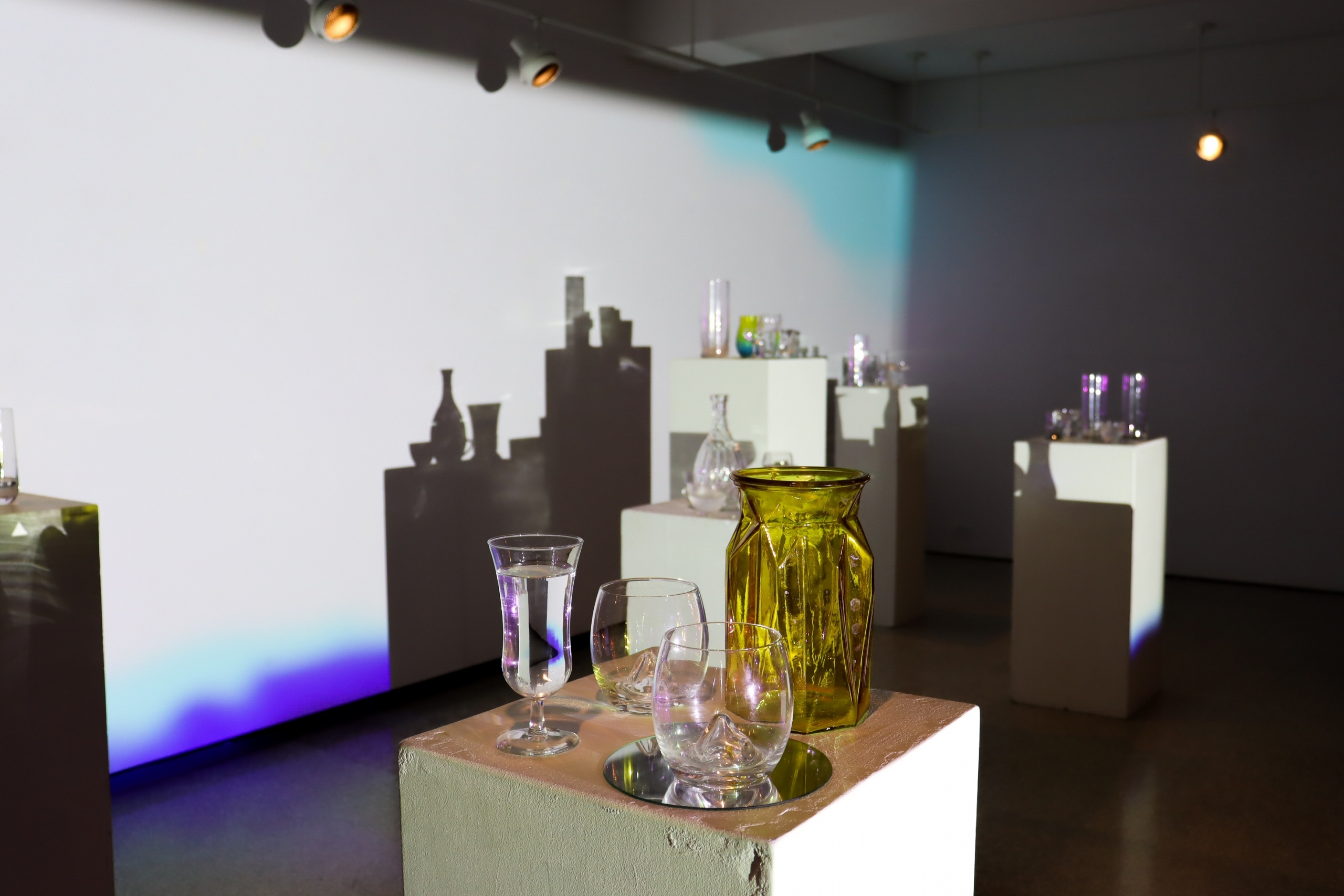 Close view of glassware and an amber vase on a plinth as sharp shadows and cool-toned projection stretch across the gallery wall.