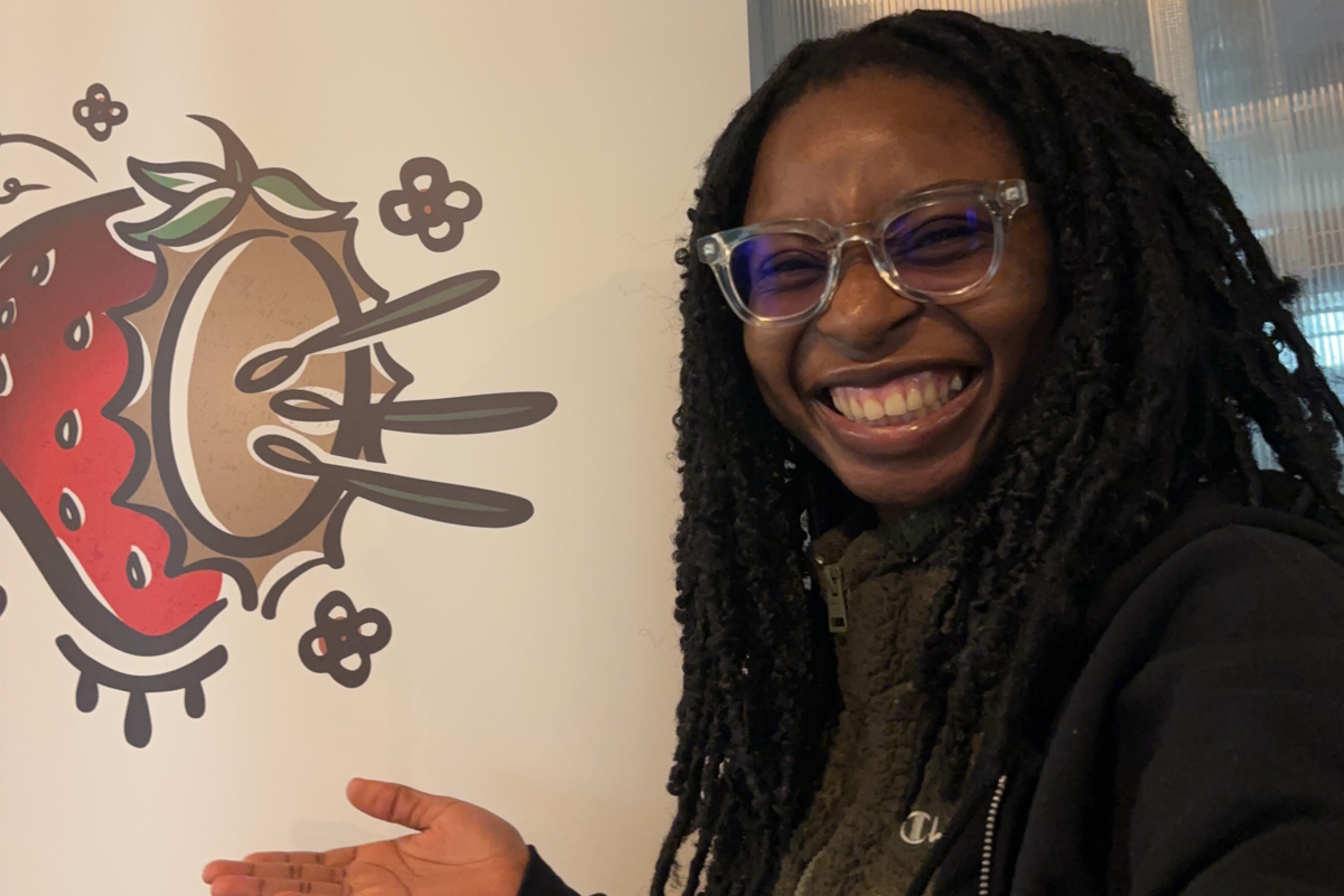 A person smiles at the camera and motions to Zoongizi Ode's logo.