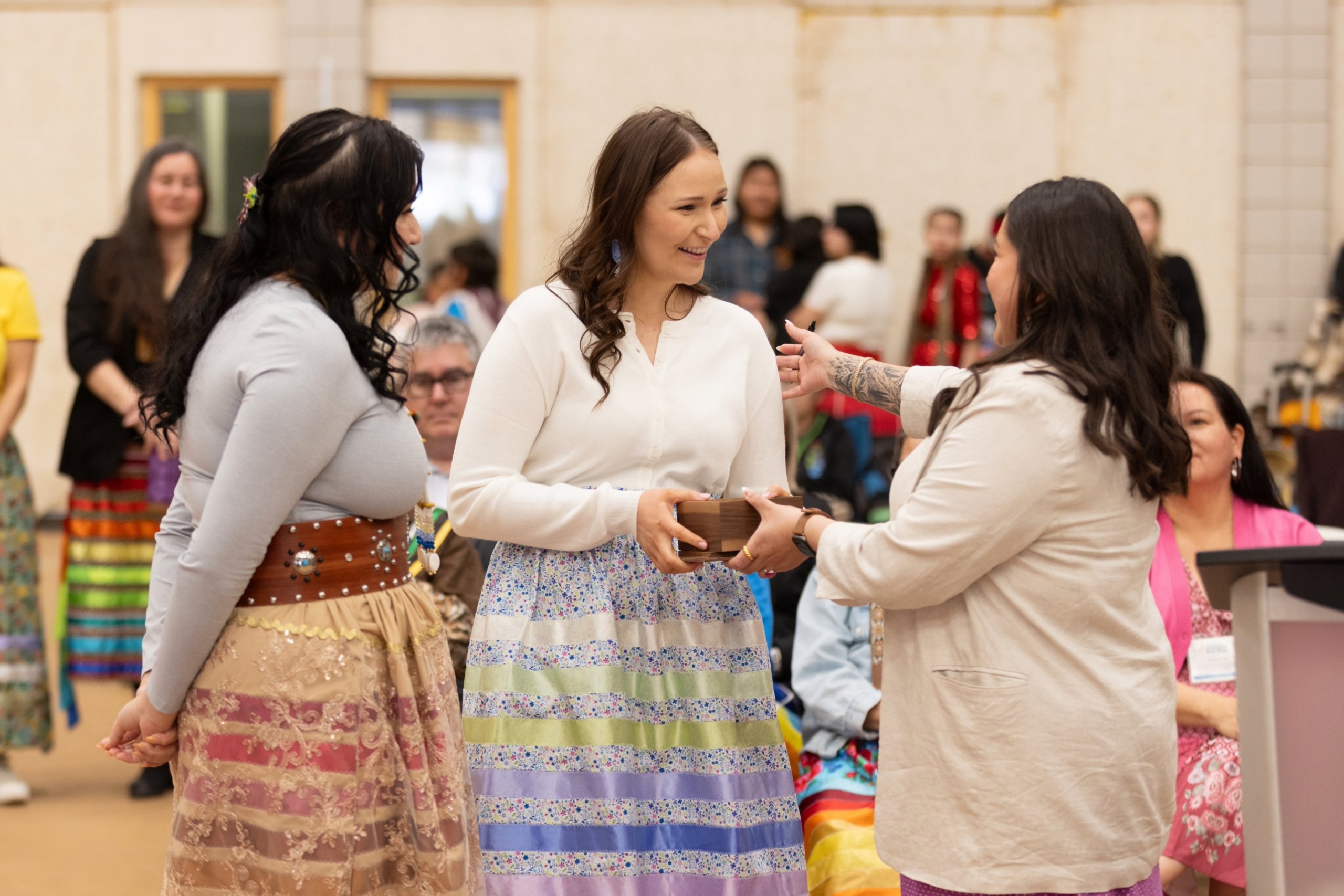 A woman wearing a ribbon skirt is handed an award from a woman wearing a blazer as a third woman looks on.