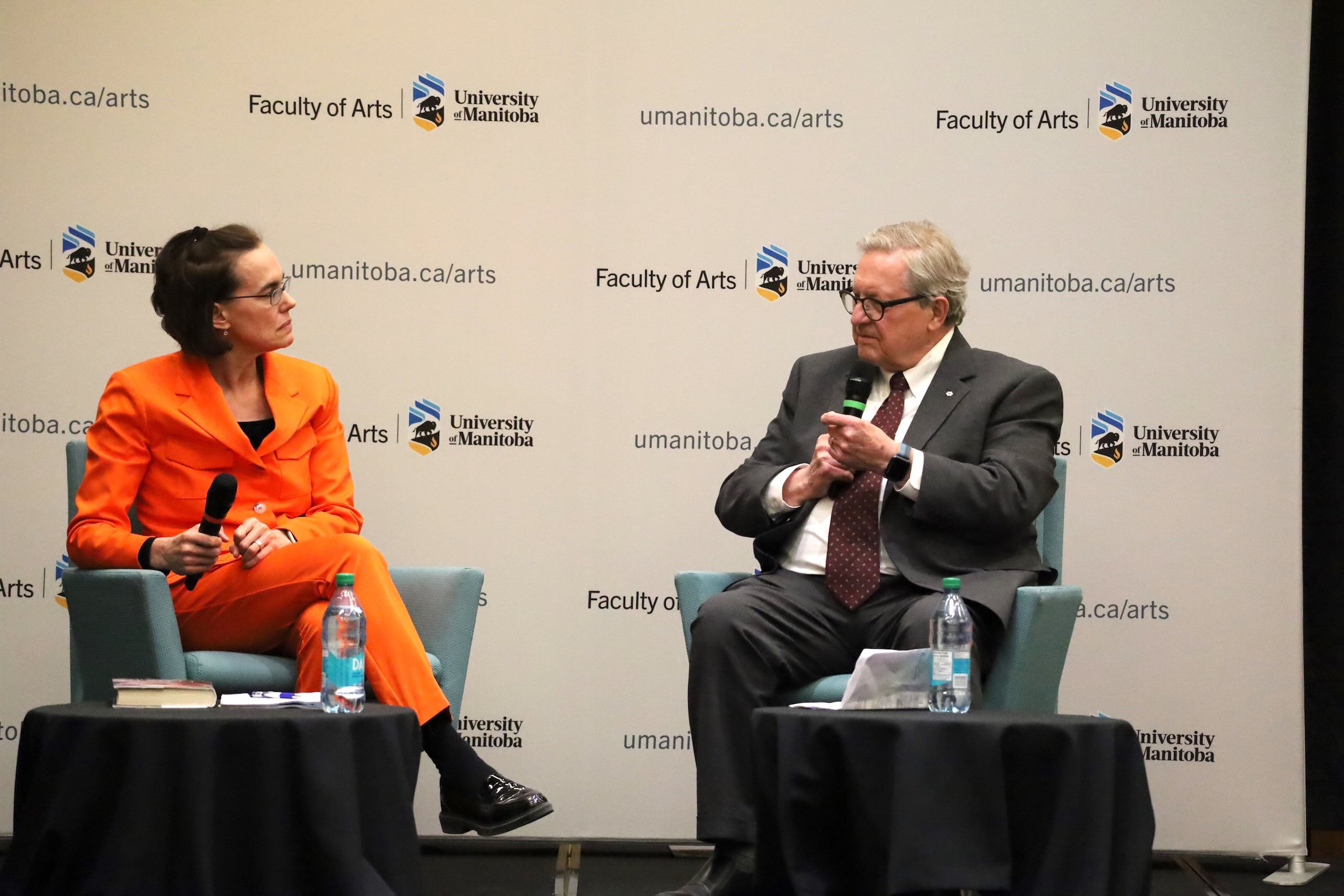 Seated woman in pale orange suit speaks with older man in glasses and suit at conference panel.
