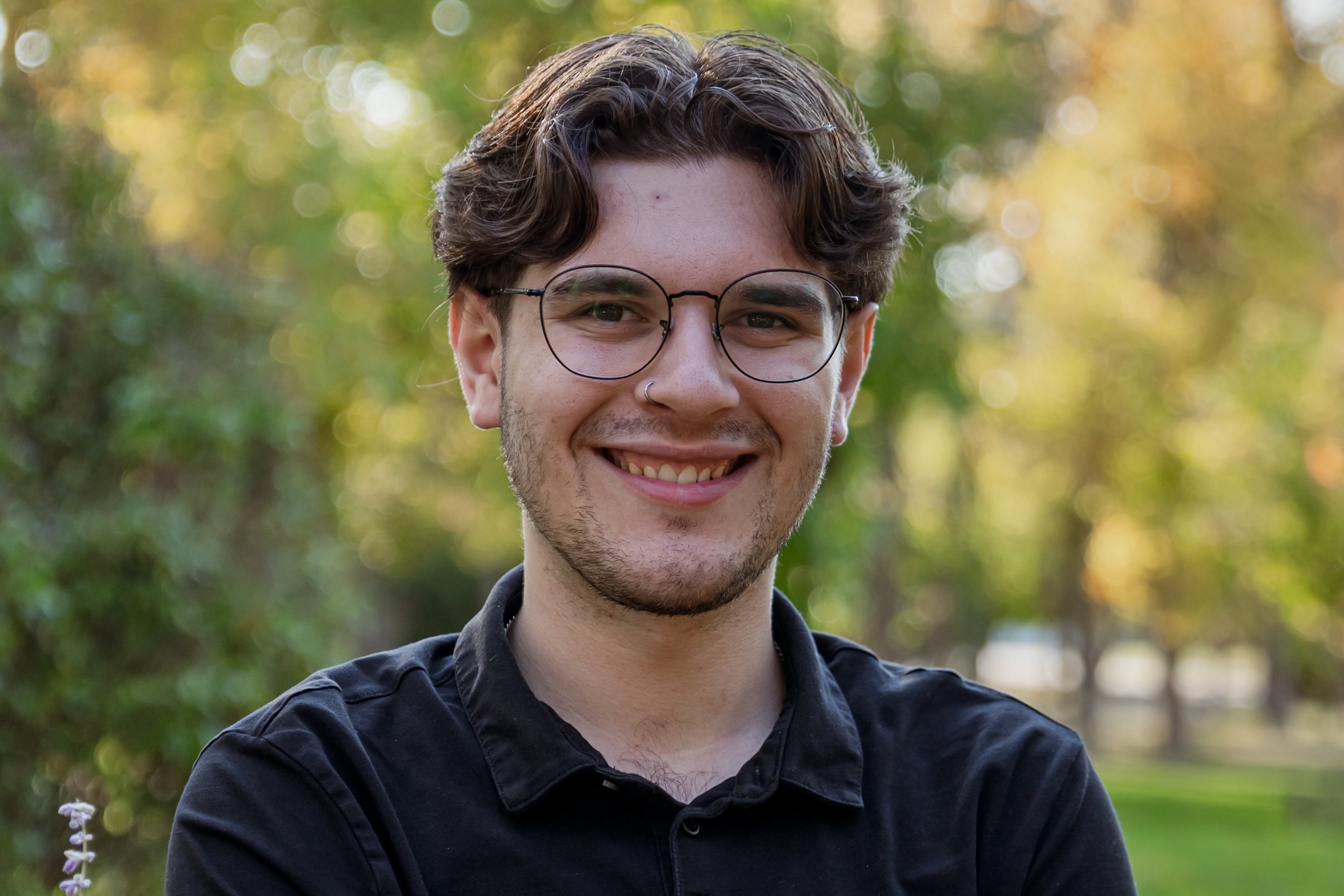 Young man in black t-shirt smiling with background of green and yellow trees.