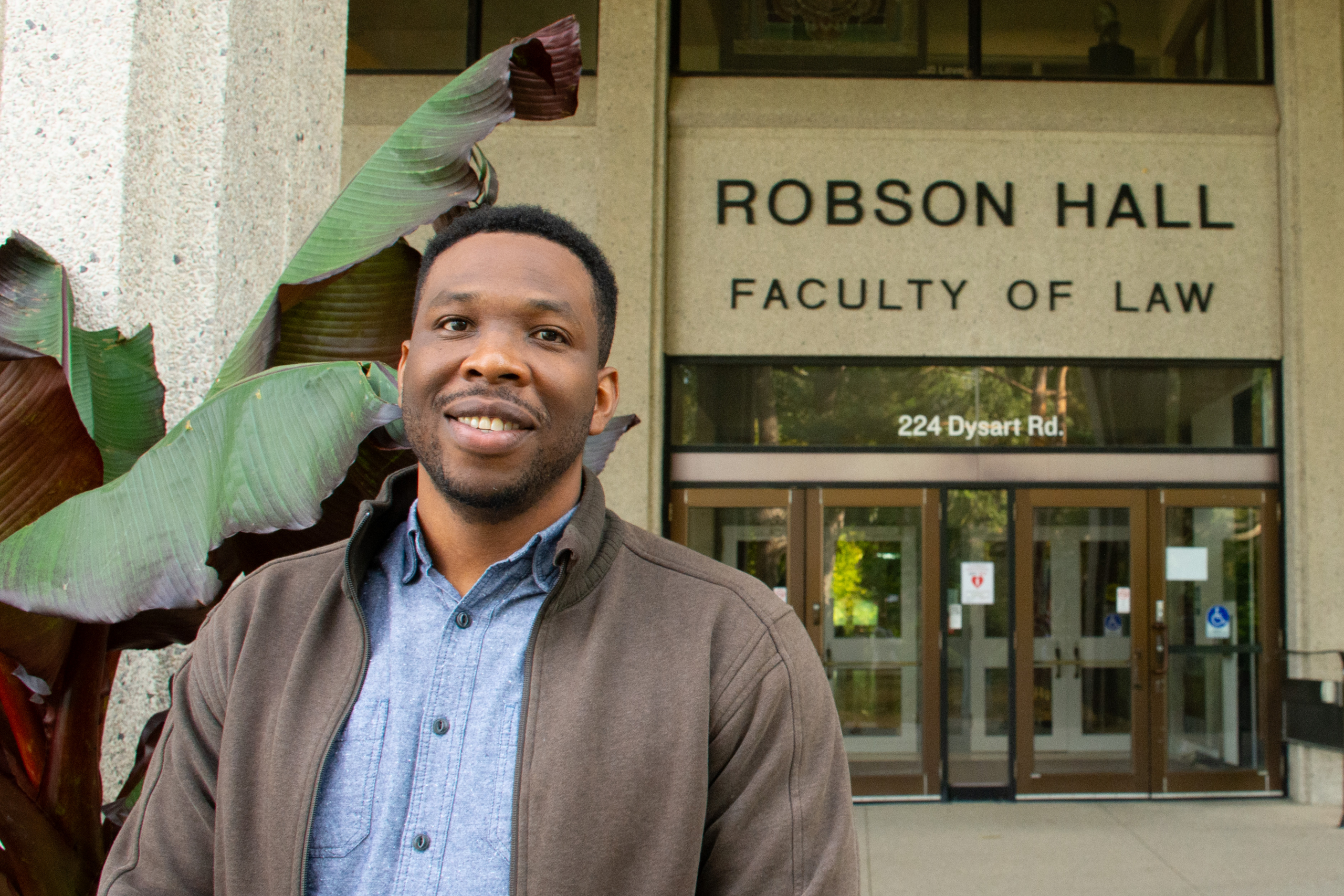 A smiling student stands next to a leafy plant in front of Robson Hall Faculty of Law front doors