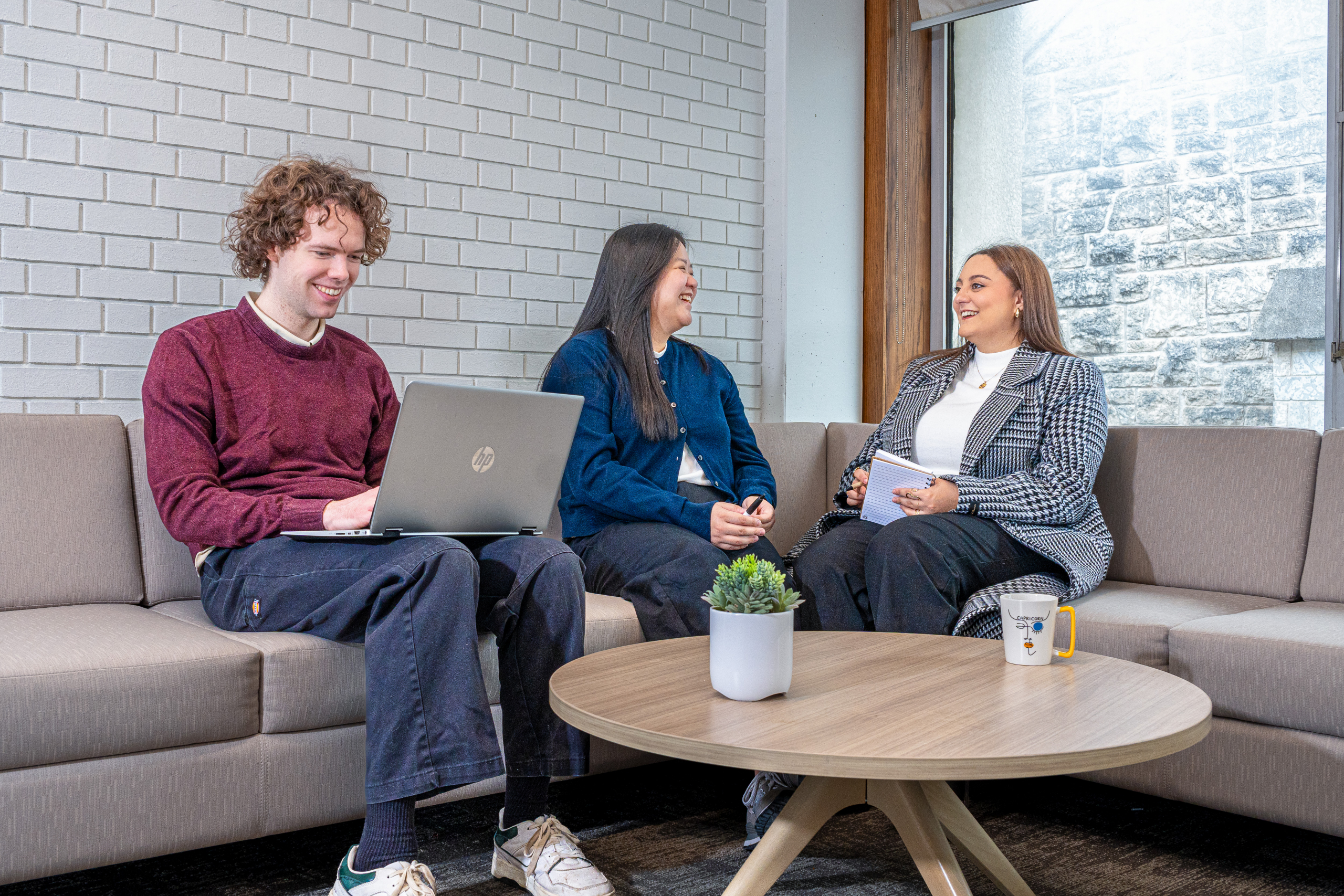 Three students sitting on a sofa in a professional environment. All wearing employee badge lanyards. One typing on a laptop. 