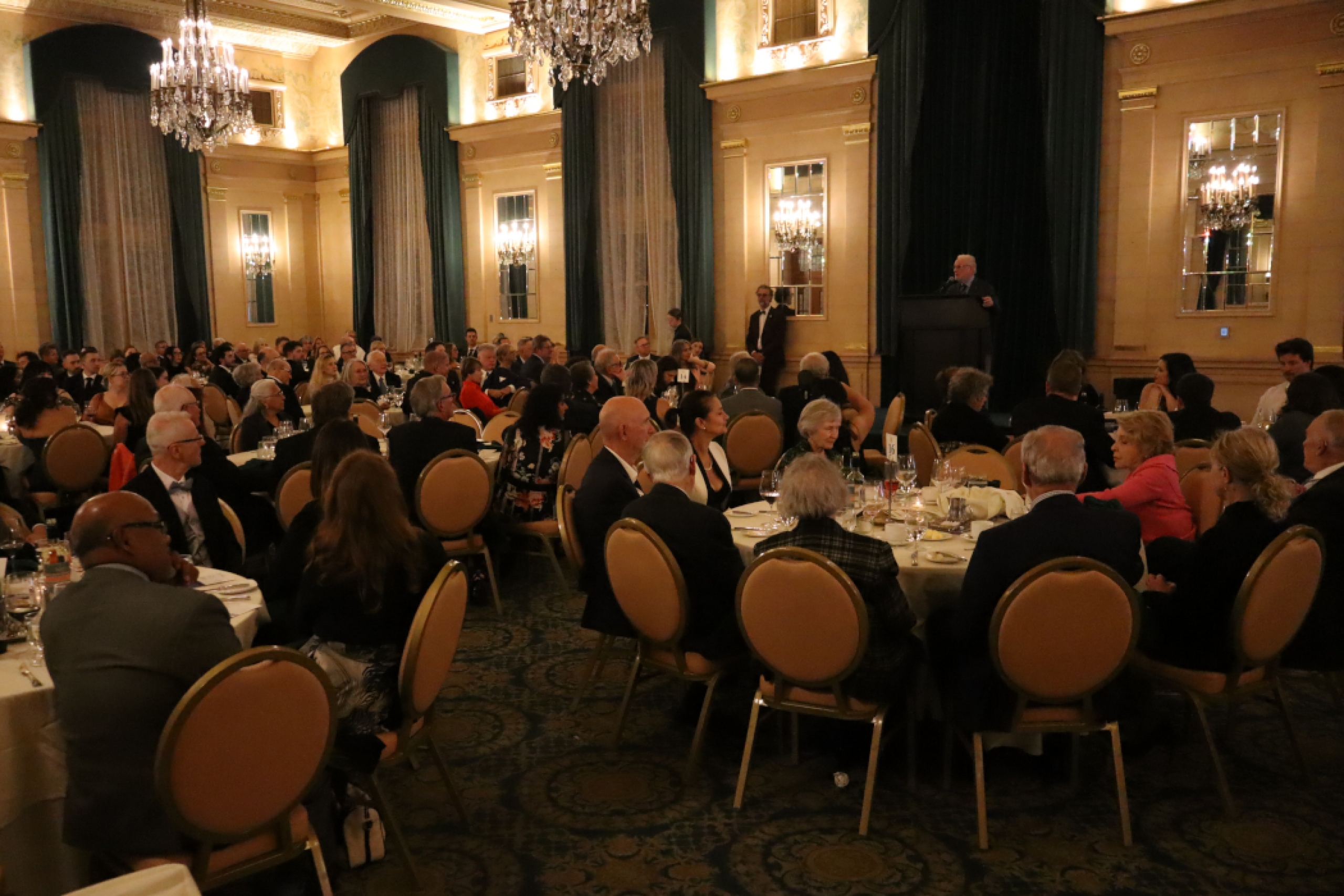 A person speaks at a lectern on a stage before a seated crowd in a ballroom.