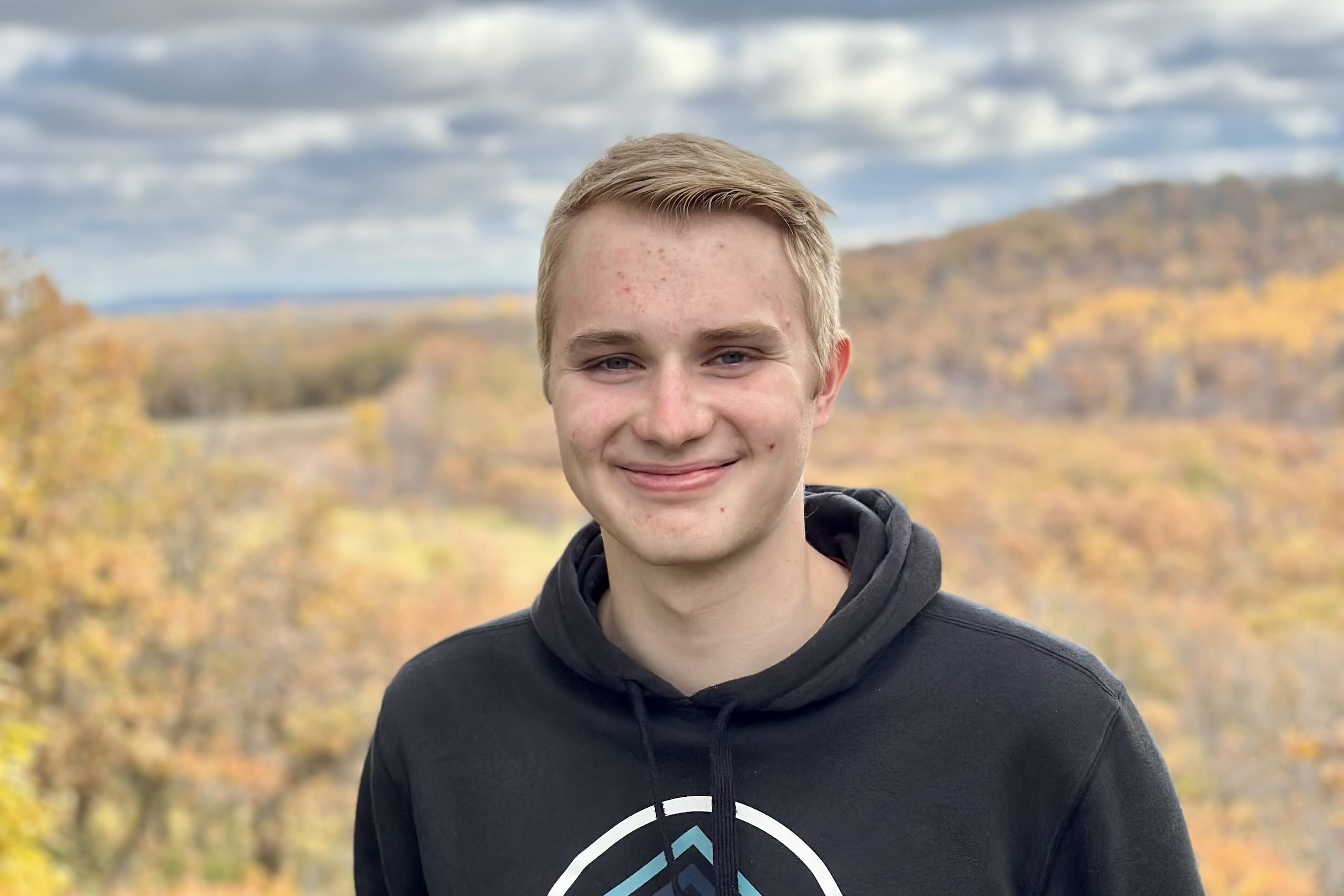 Headshot of smiling young man in black hoodie outdoors with hills and fall foliage behind him.