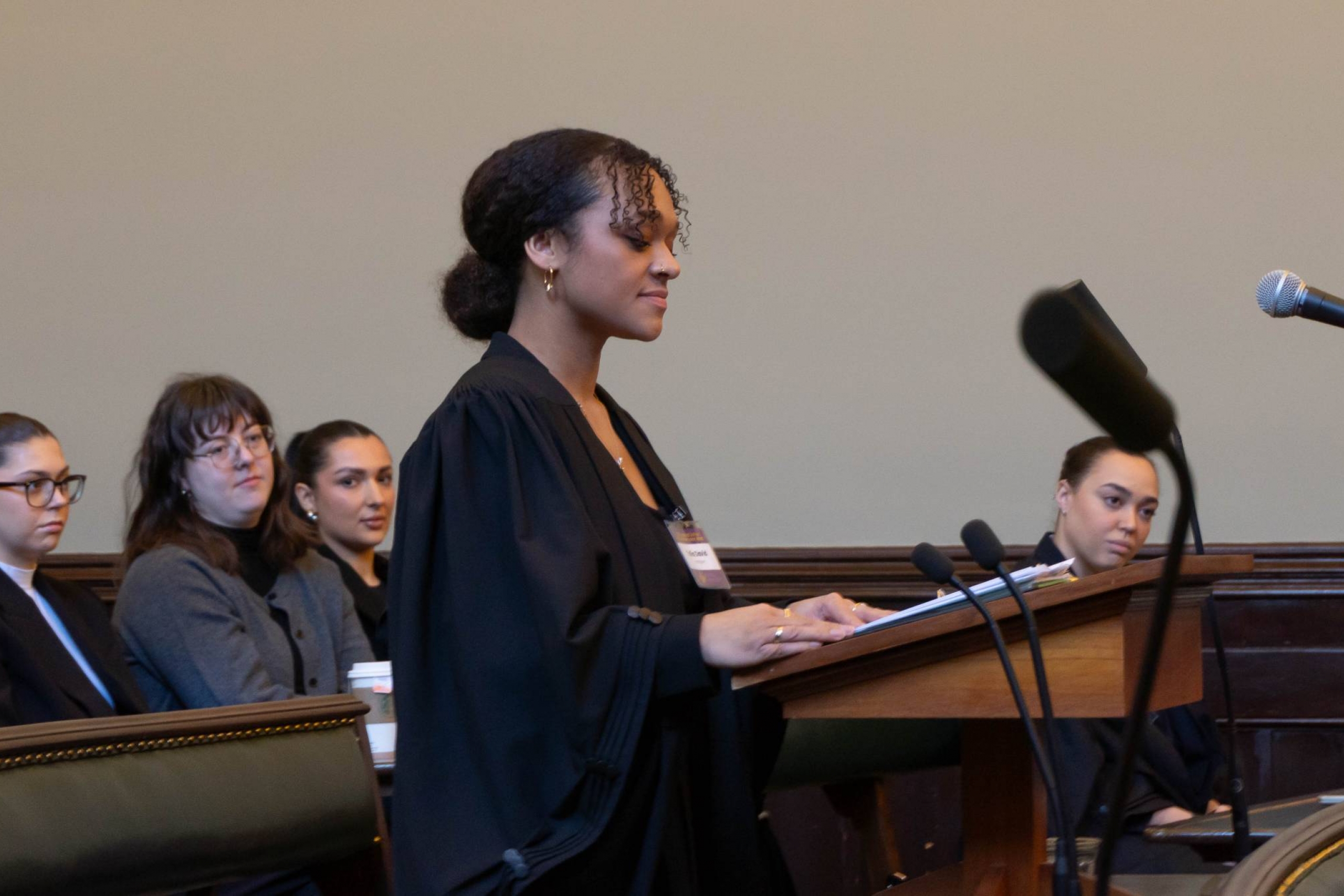 A young woman with black curly hair in a pony tail in a black lawyer's robe speaks at a podium while her teammate listens. 