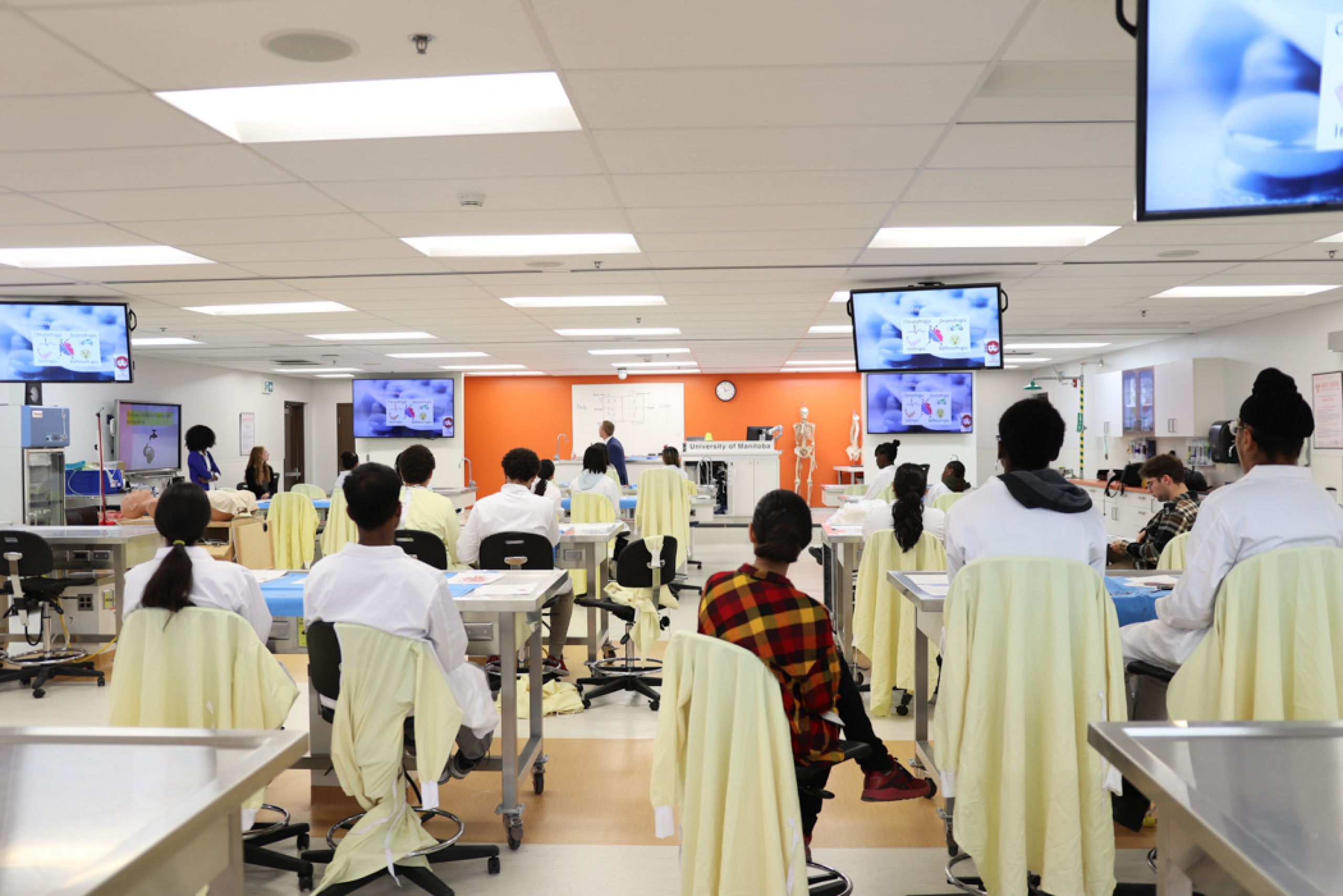 A wide lab shot taken from the back of the room shows campers with their backs to the camera, listening to a clinical lecture. Five ceiling-mounted TVs display the same visual.