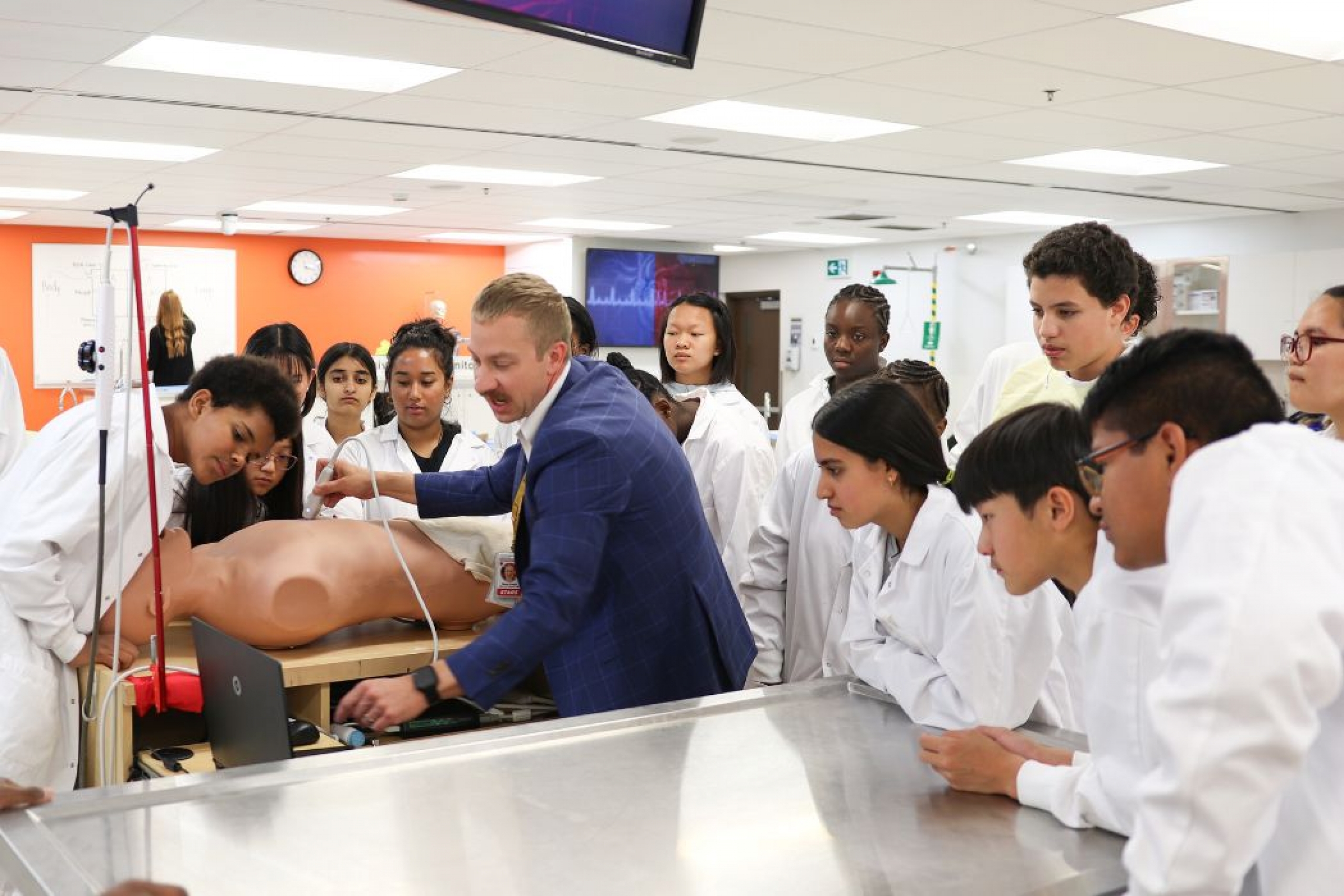 Danny Kustra, clinical education coordinator at UM Rady Faculty of Health Sciences, holds an ultrasound probe to a manikin’s abdomen during a simulation. A group of campers leans in to watch the laptop monitor displaying the ultrasound image.