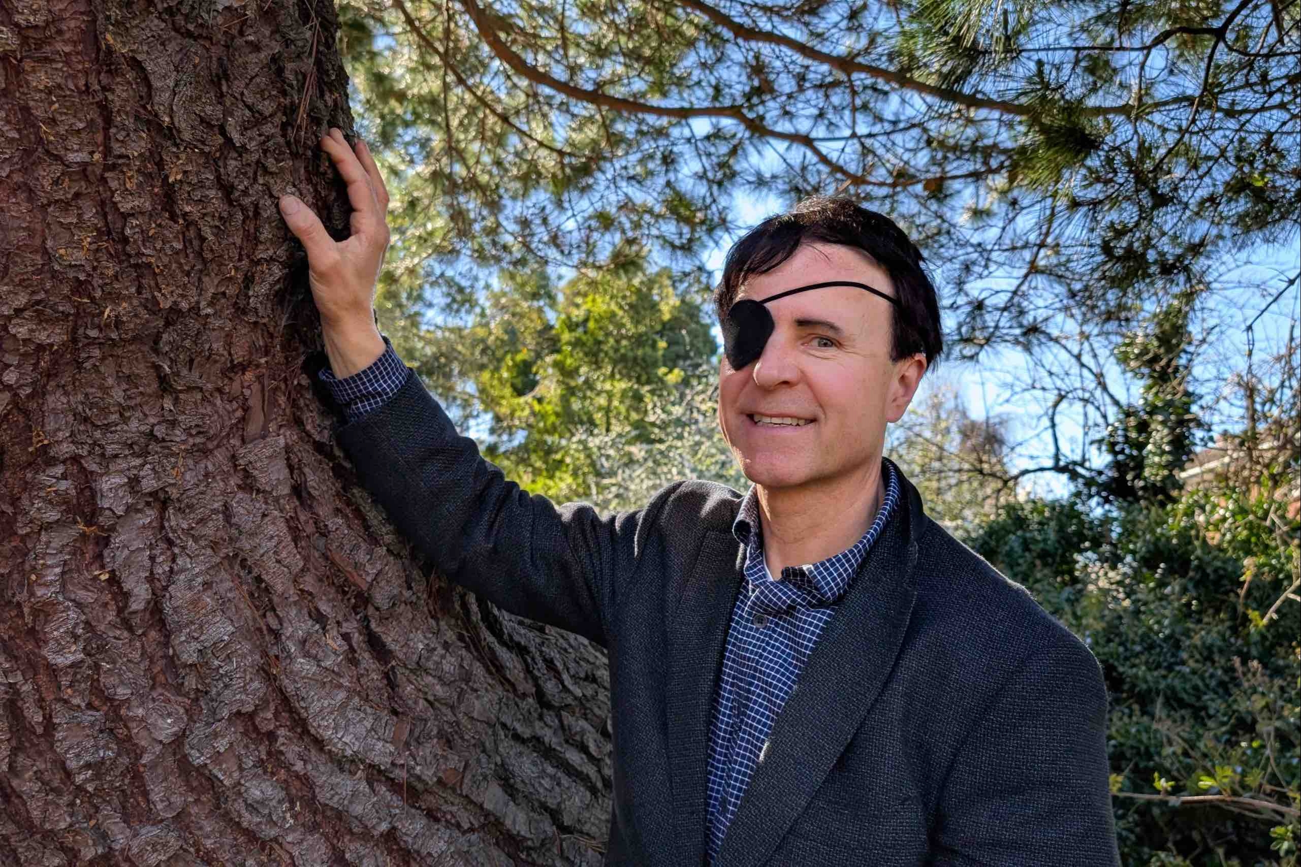 a smiling man in blue suit jacket rests an arm against a large tree