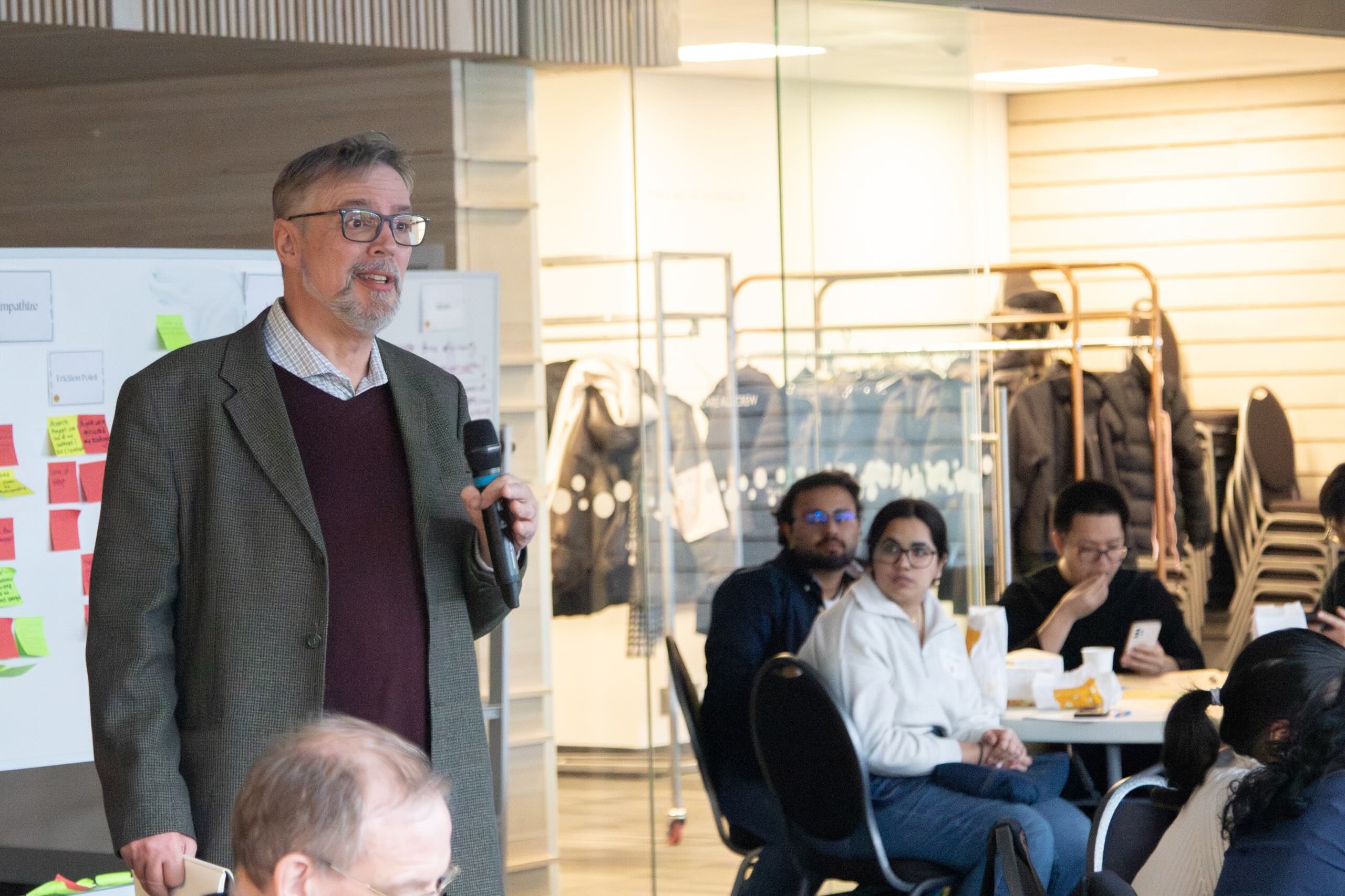 Standing man with microphone addressing crowd seated at round tables.