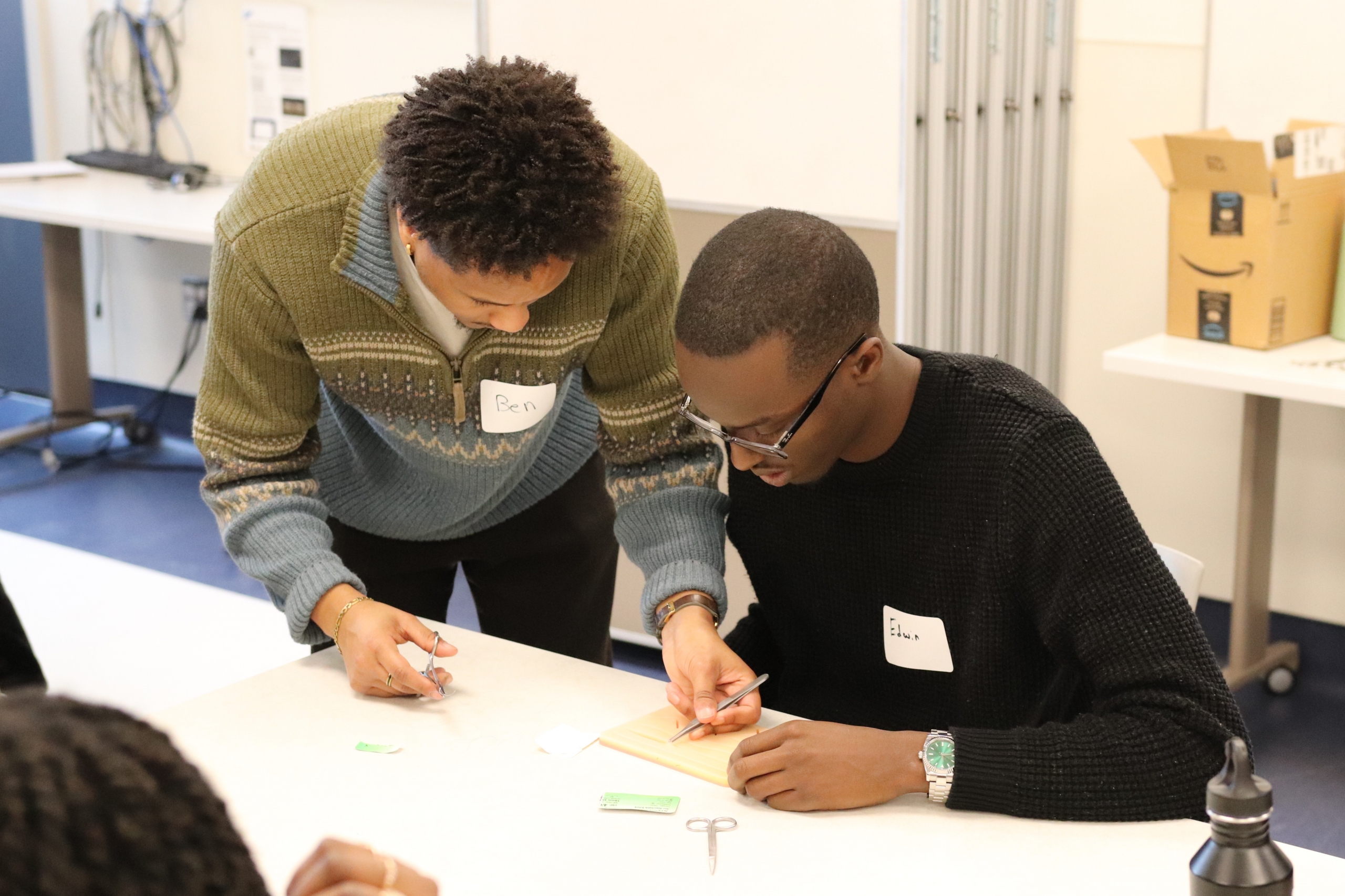 The medical student is holding scissors in one hand and tweezers in the other. He is showing the undergraduate student how to suture.