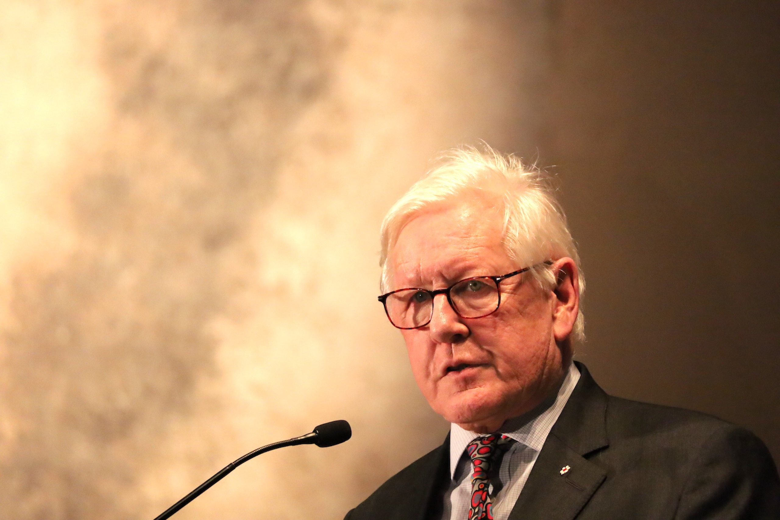 Older man with white hair and glasses speaks into a black microphone.