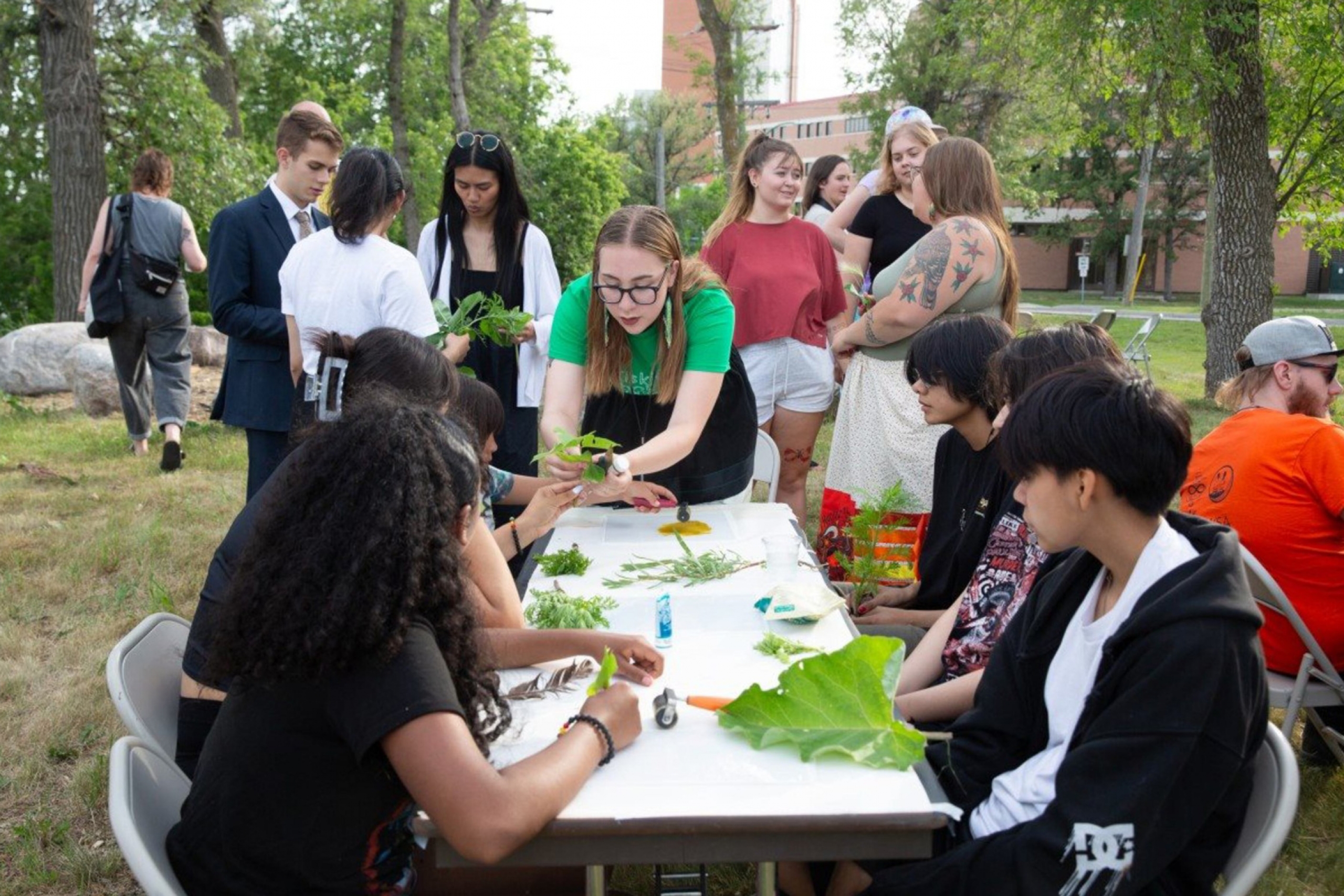 Jory Thomas-Blanchard with student looking at various flora around a table on the south green space on the river bank across from the Asper School of Business.