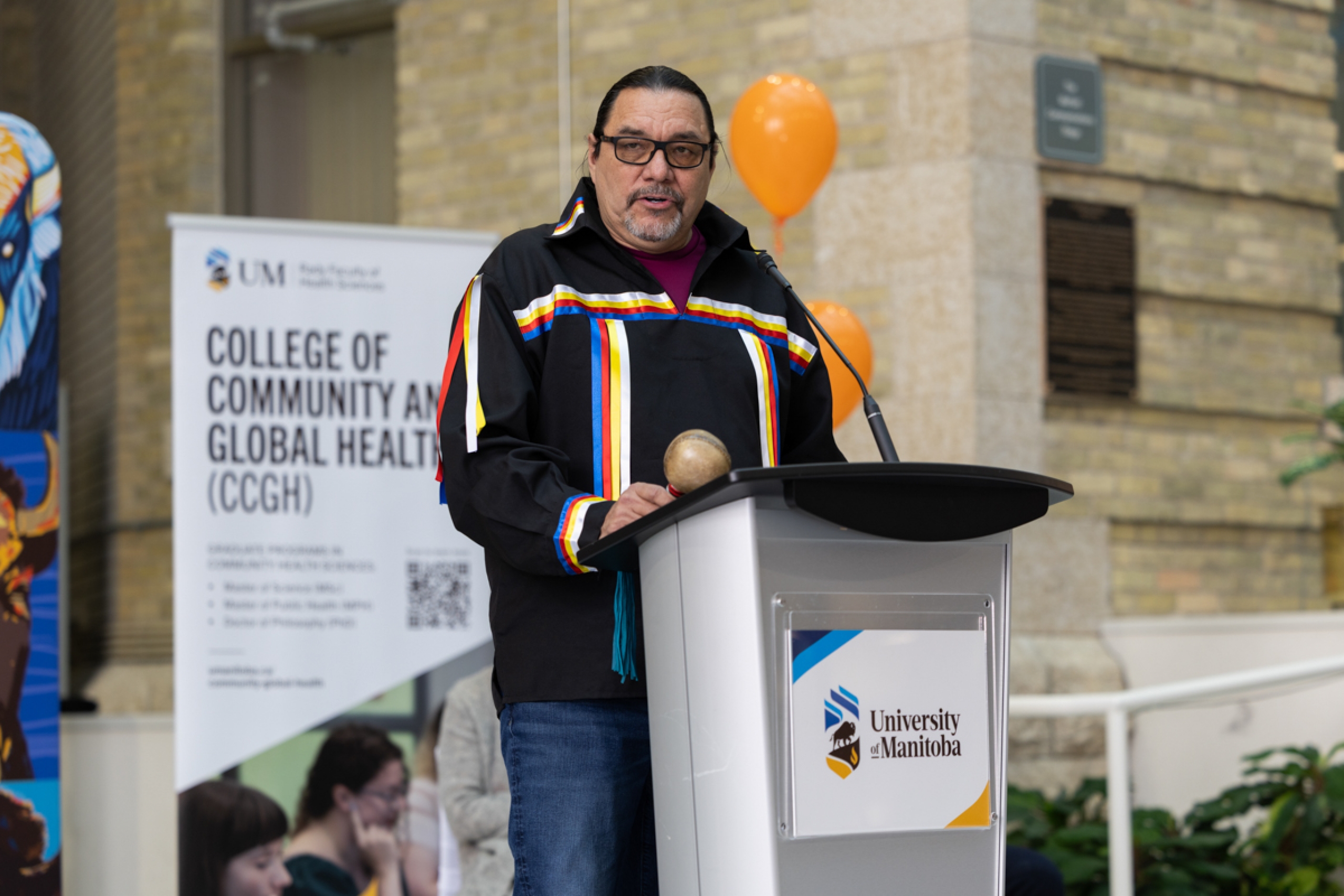 Elder George speaks at a podium onstage. The stage is decorated with balloons and university-branded banner backdrops.