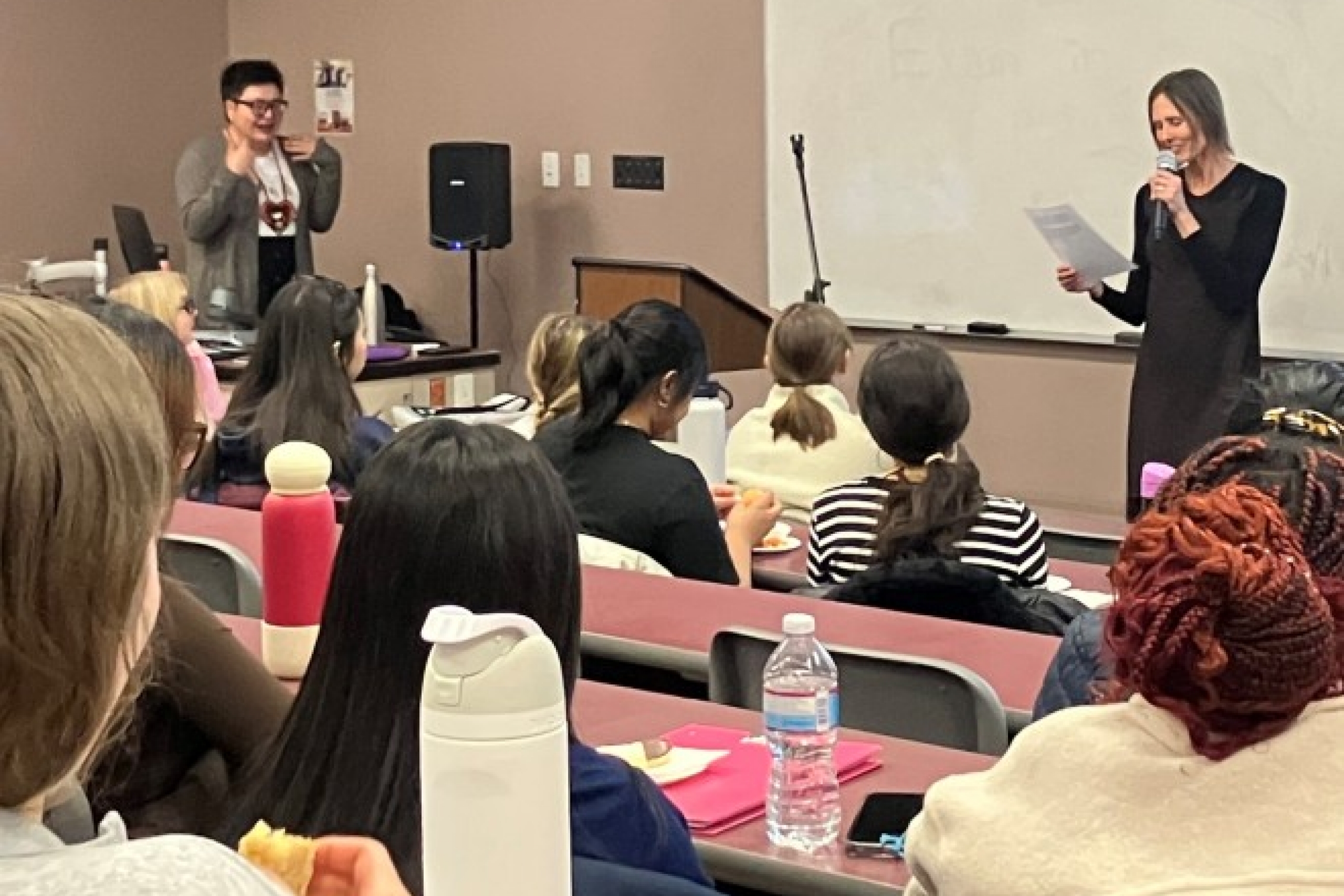 Two women presenting at the front of a classroom full of students.