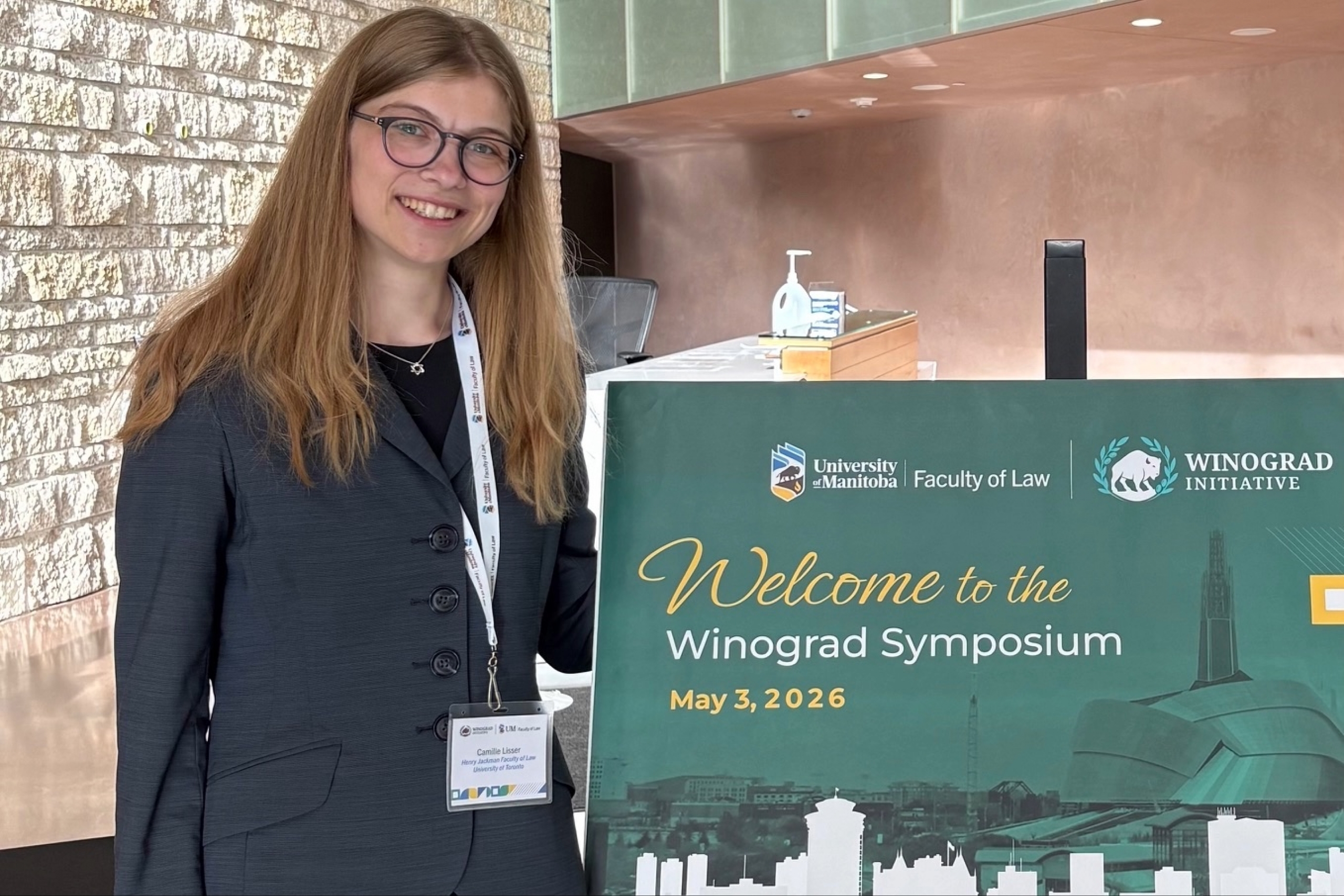 A student in a suit jacket smiles beside the Winograd Symposium sign