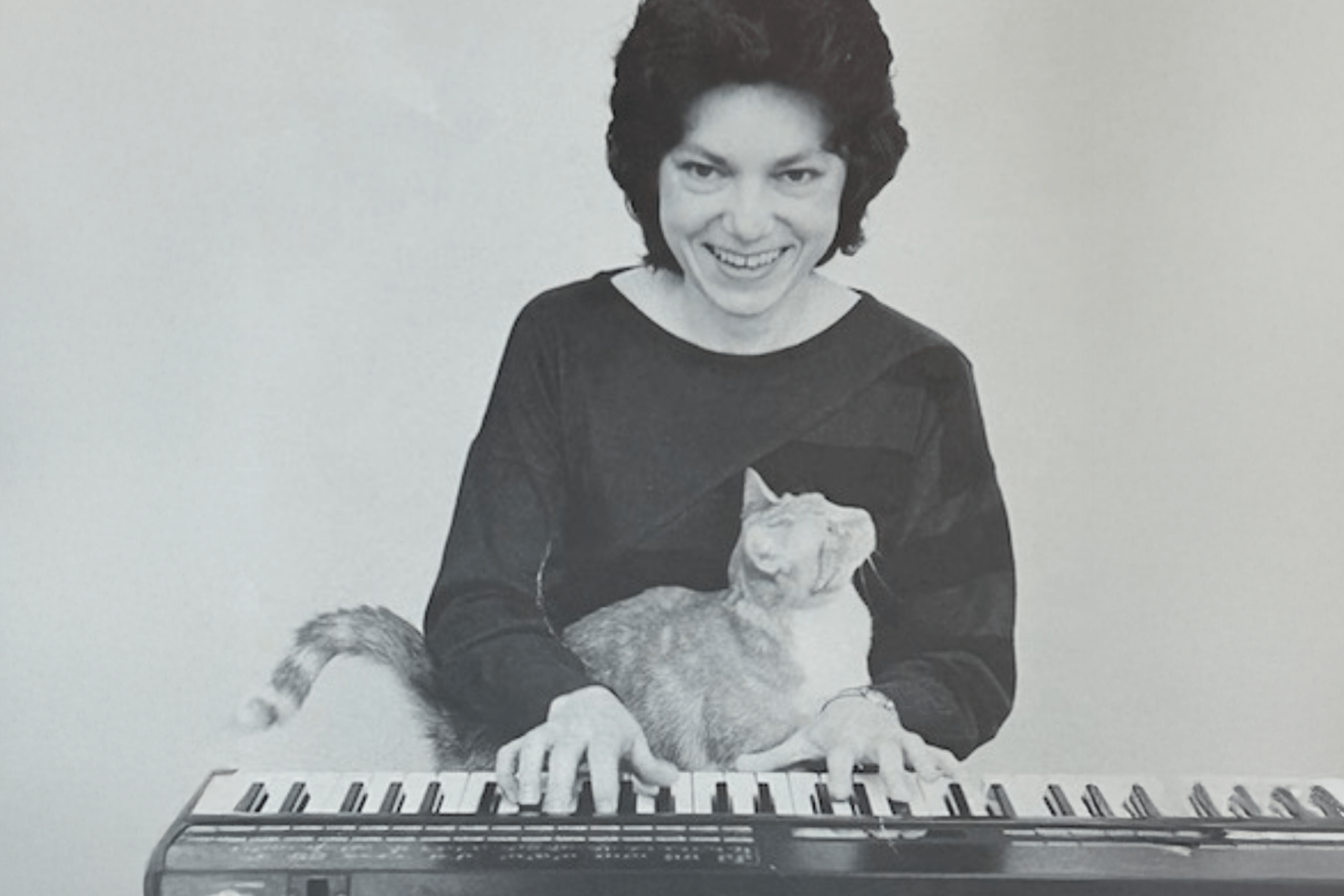 Black-and-white photograph of a smiling woman seated at an electronic keyboard while a cat sits across the keys in front of her.