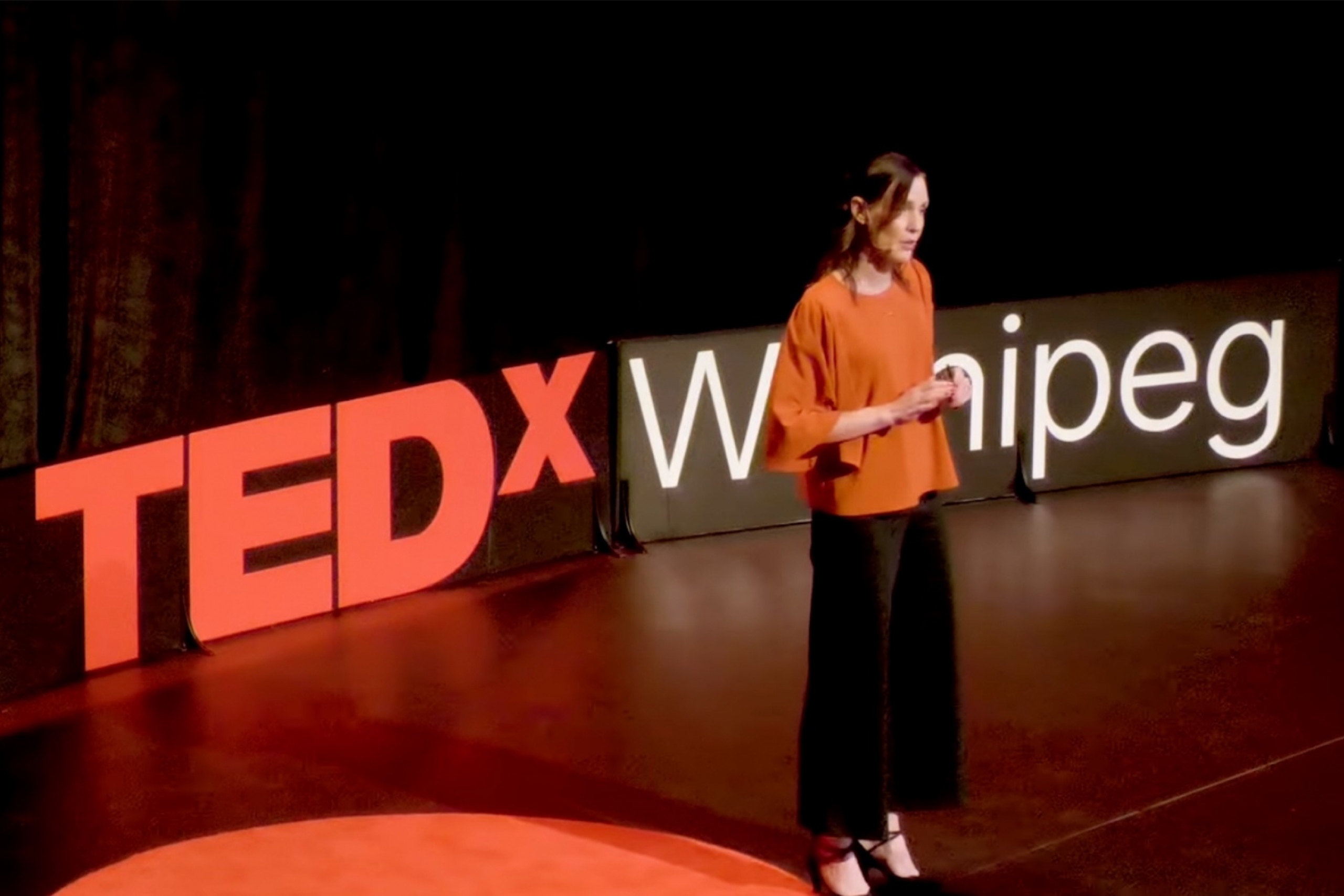 Spotlighted woman in orange top on dark stage with red letters spelling Tedx Winnipeg.