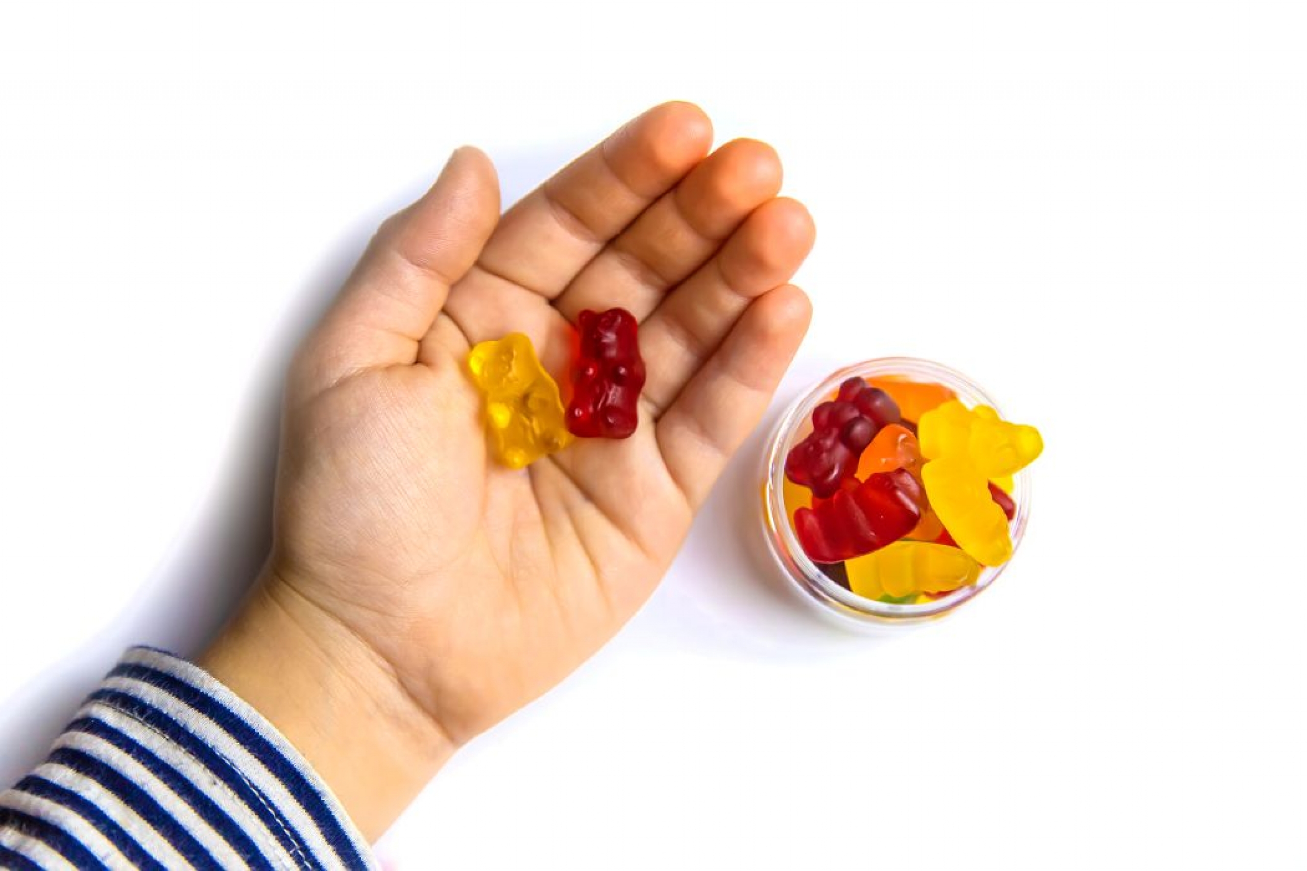 A bowl of gummy bears sits beside a child's hand holding two gummy bears.