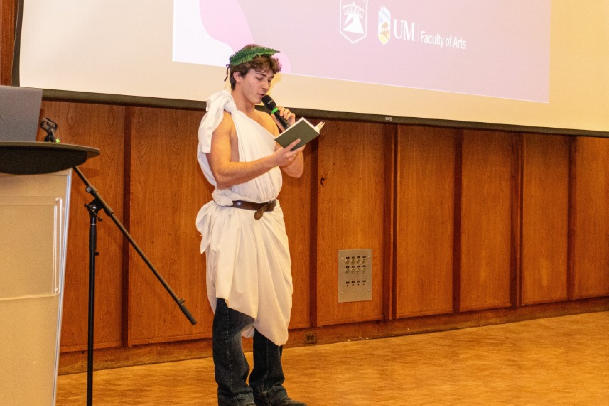 A student wearing a toga and a laurel head wreath reads a poem from a book.