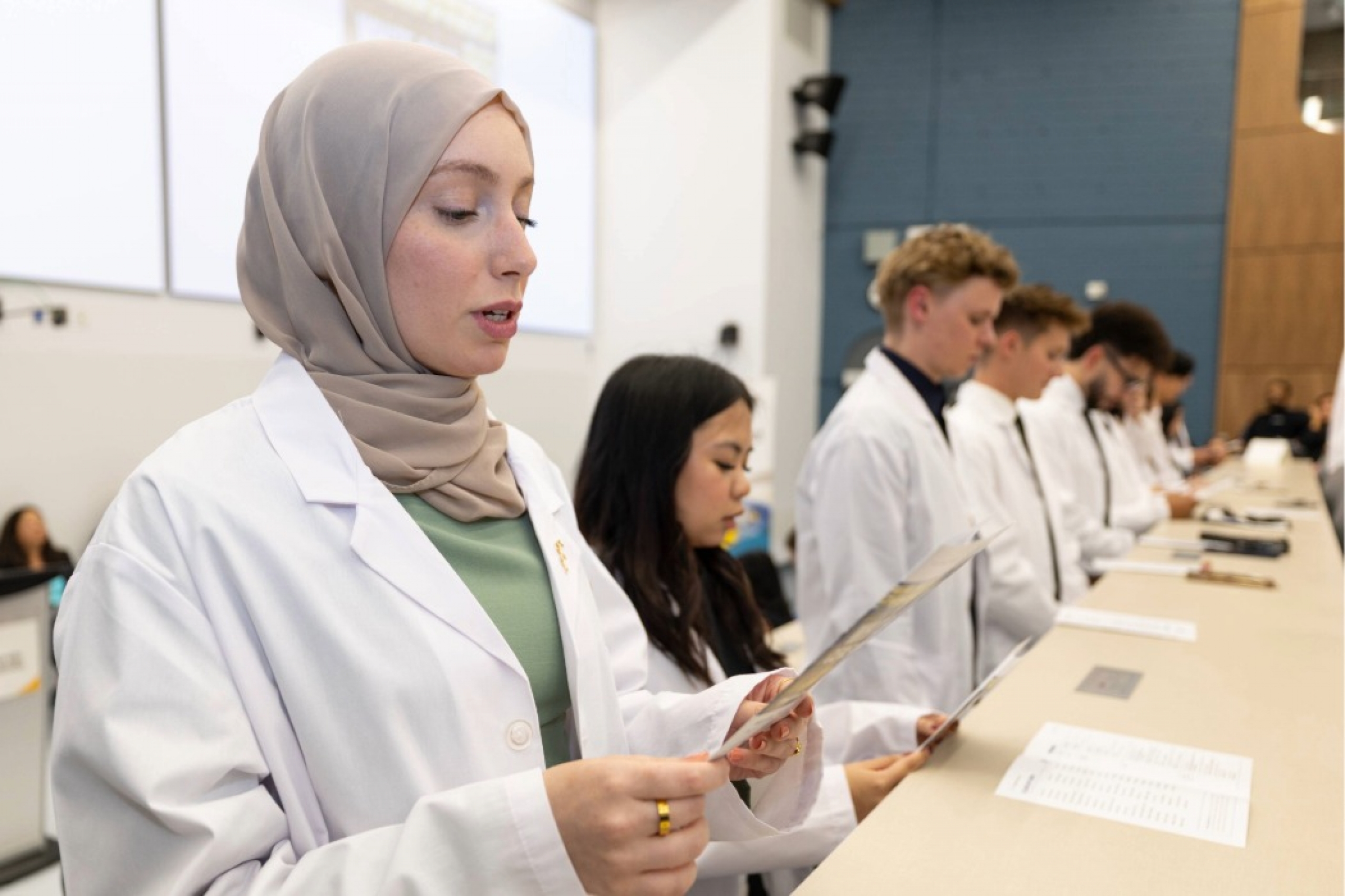 Pharmacy students standing in a row, reading from a program as they recite their pledge of professionalism.