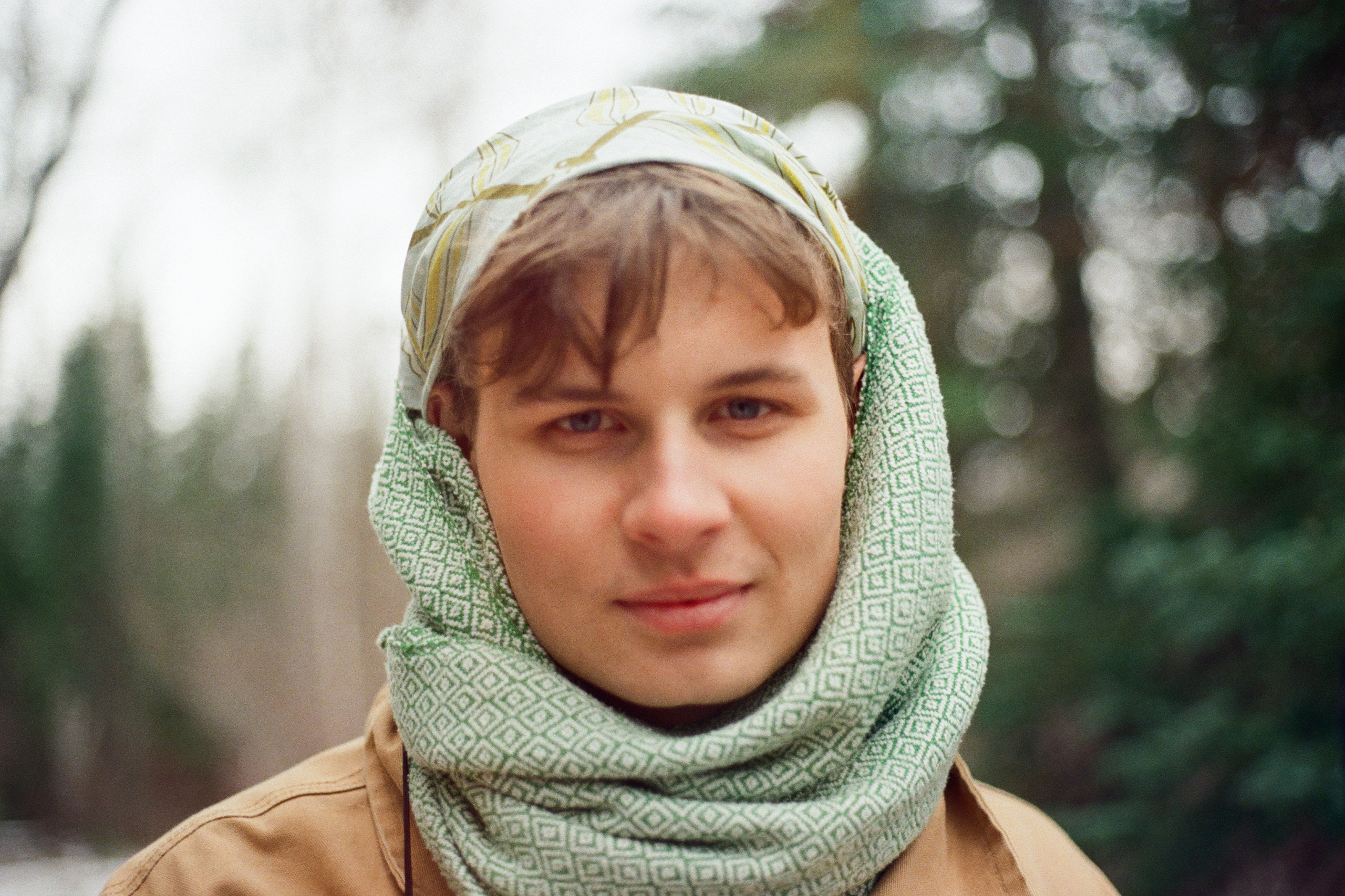 Portrait of artist Cole Osiowy outdoors, wearing a patterned green scarf and head covering, looking directly at the camera.