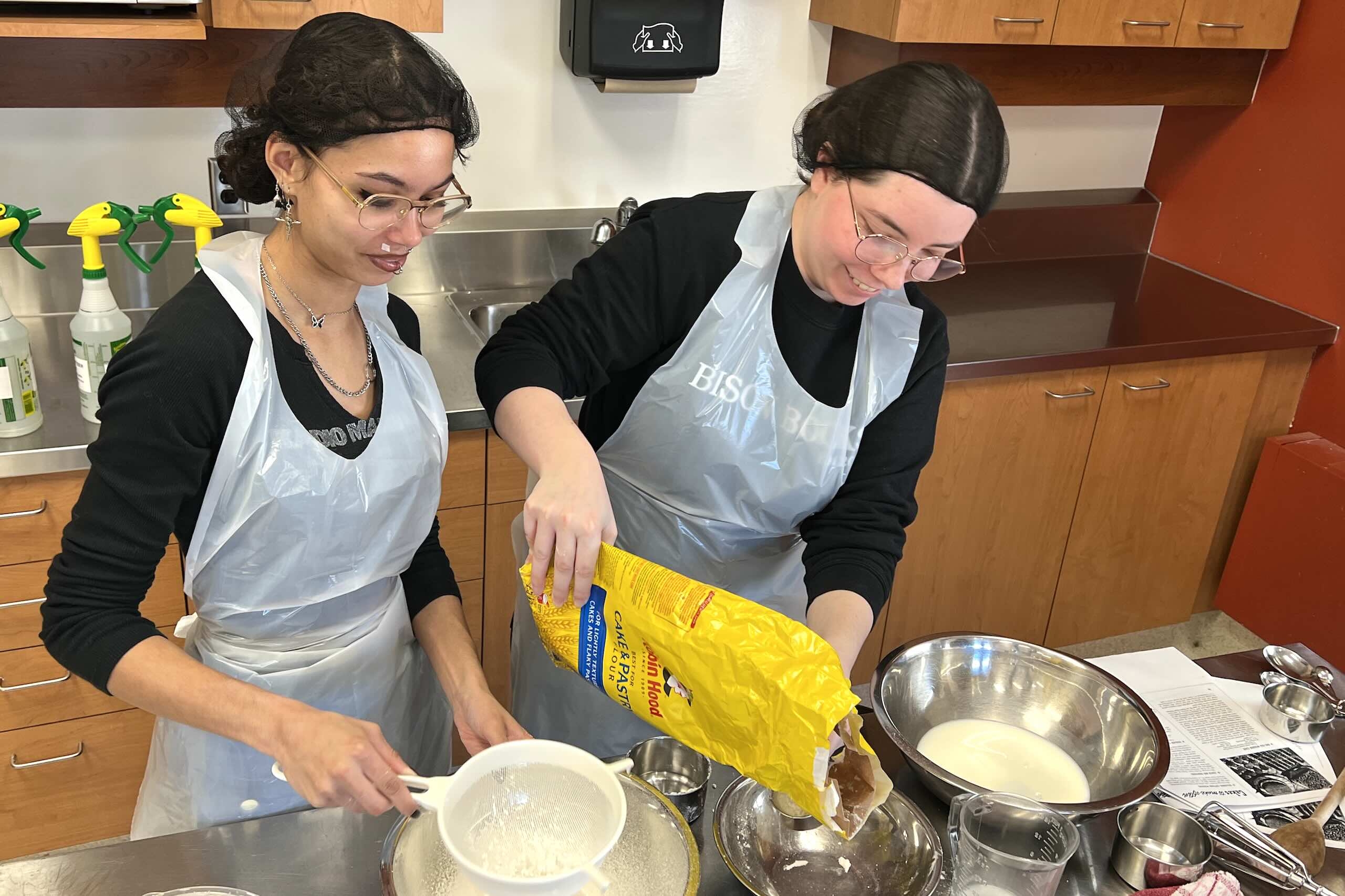 Two students with aprons and hairnets, one mixing batter, one pouring flour into a measuring cup from a yellow bag.
