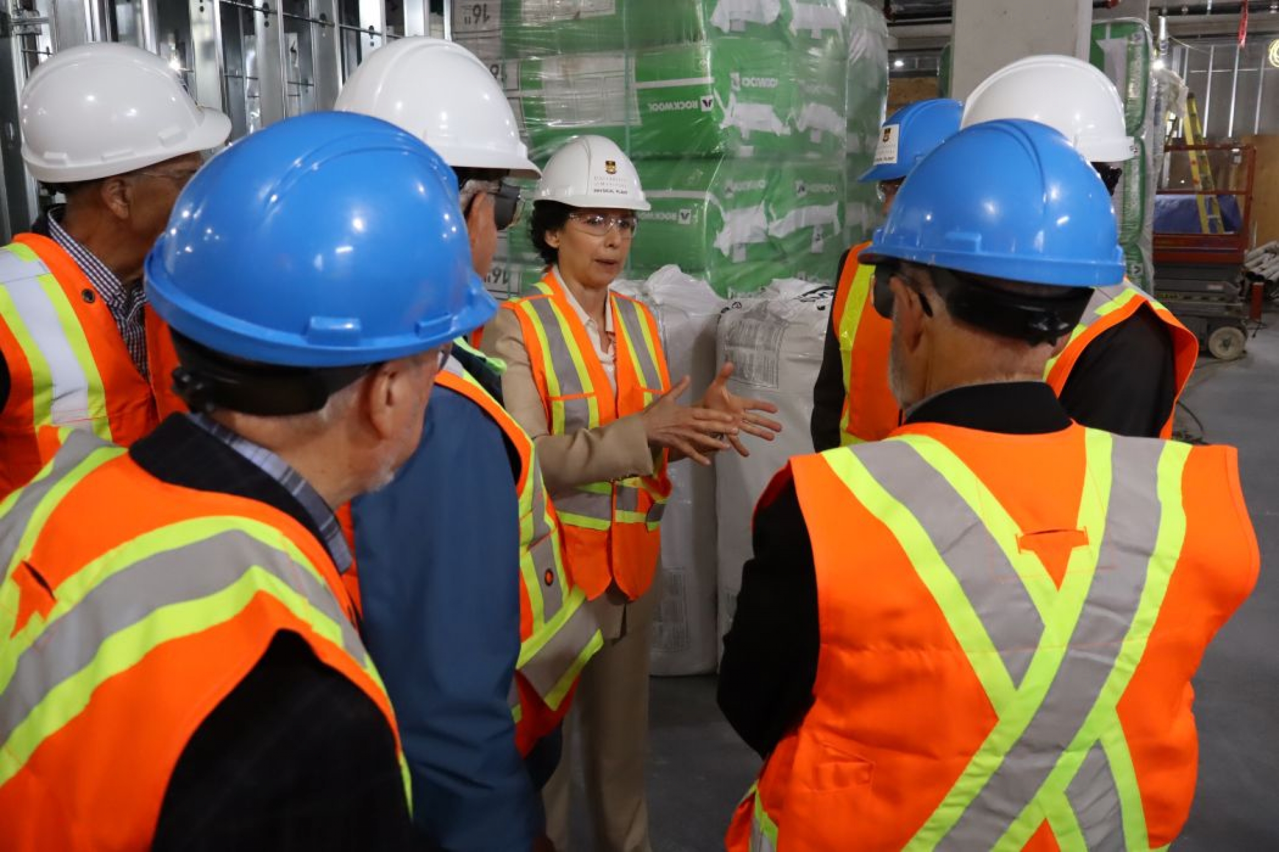 Seven people wear hard hats, safety glasses and high-vis vests. They are talking.