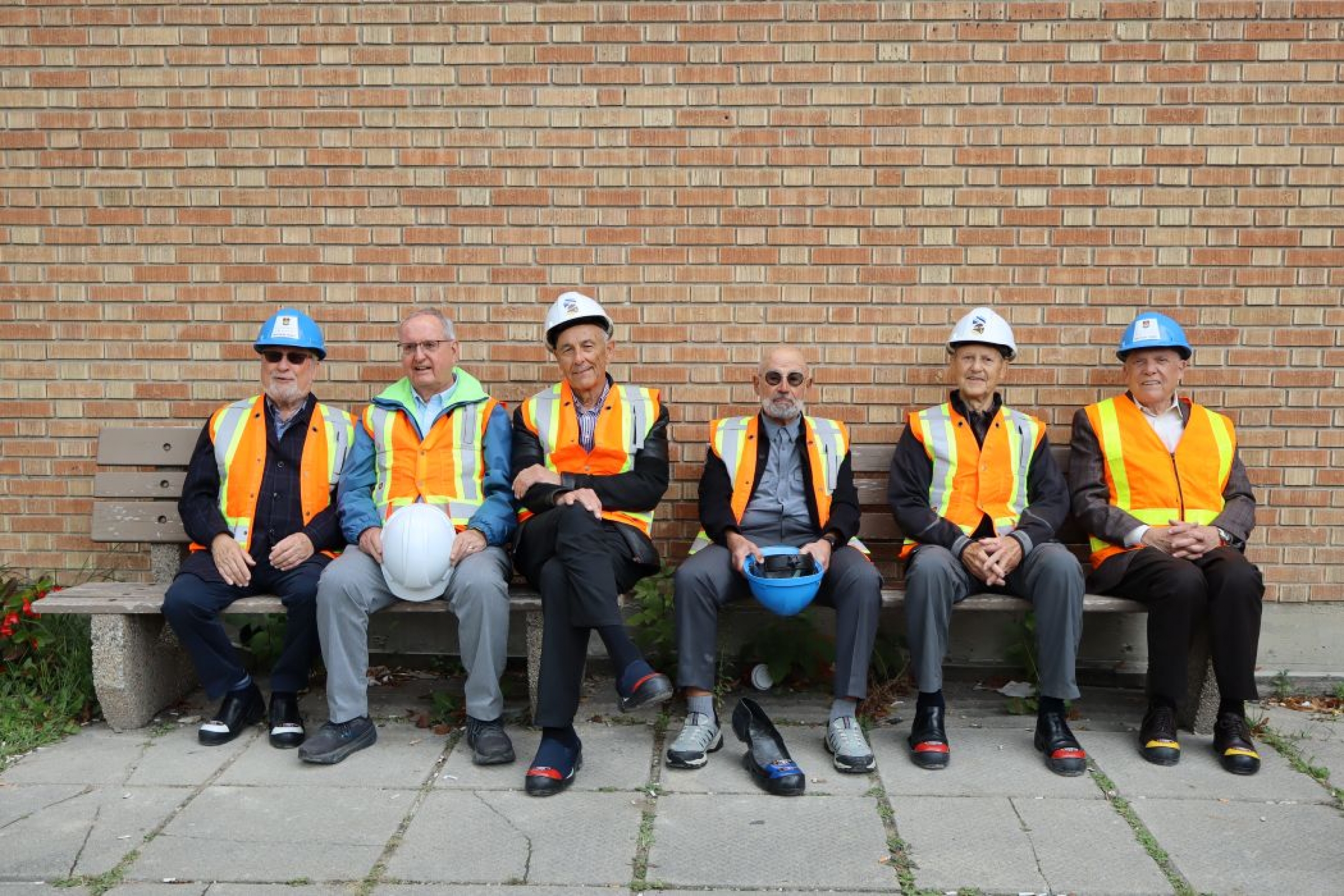 Six people sit on a bench. Some are wearing hard hats and high-vis vests. 