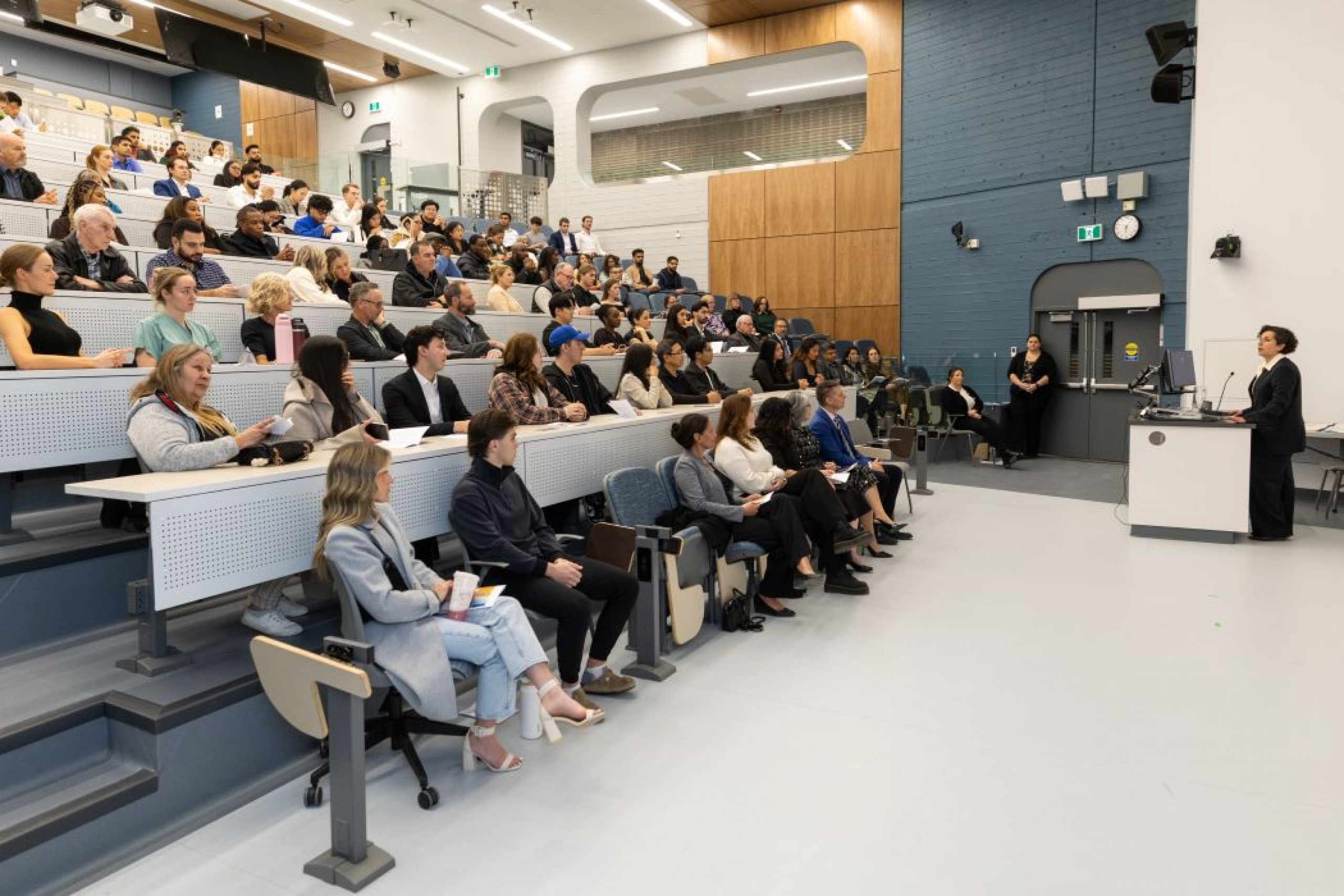 The dean speaks into a microphone at a lectern in the front of the room. The lecture hall is filled with people.