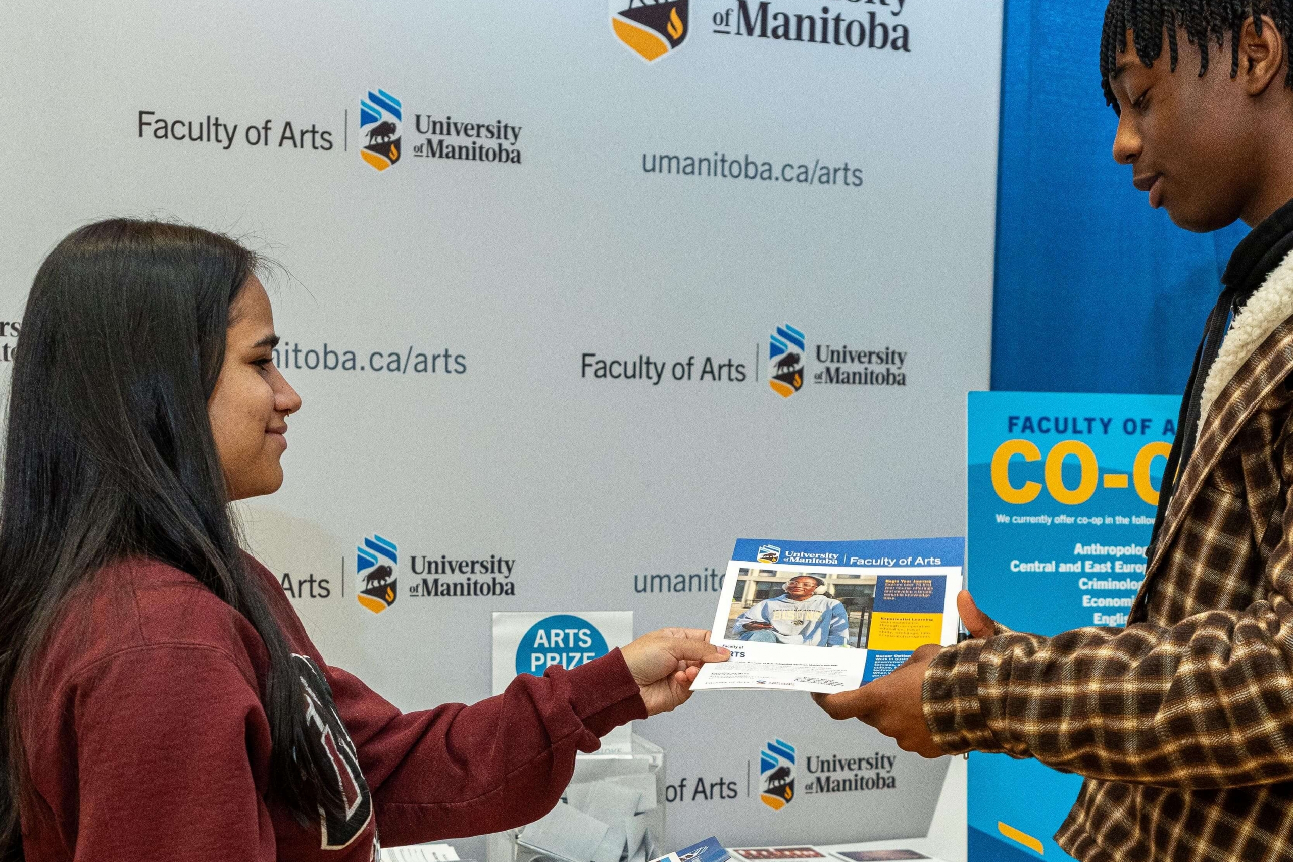 Student handing a pamphlet to another student in front of a trade show booth.