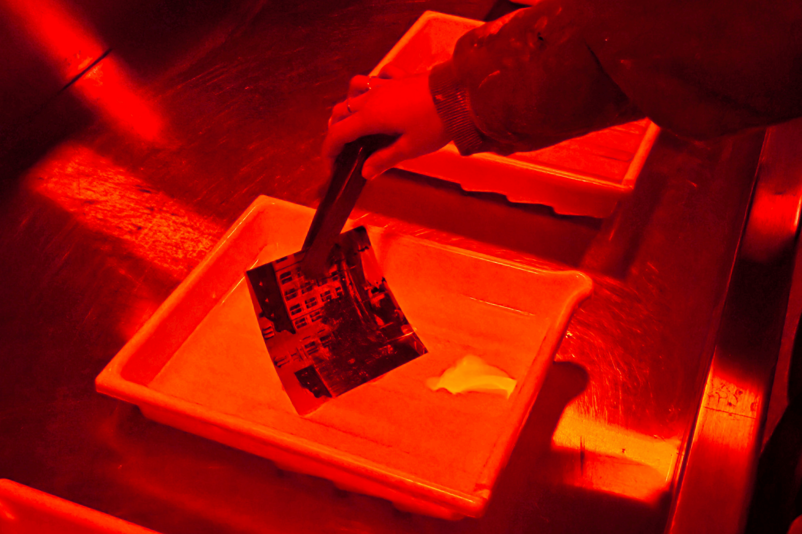 A hand uses tongs to lift a photographic print from a developing tray under red safelight in a darkroom.