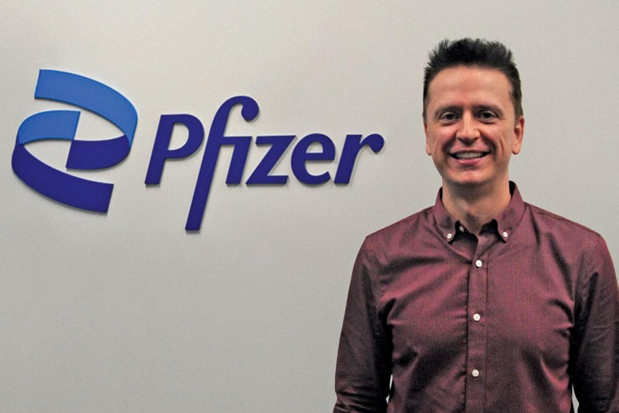 A person smiles at the camera. To the left is a Pfizer wall signage.