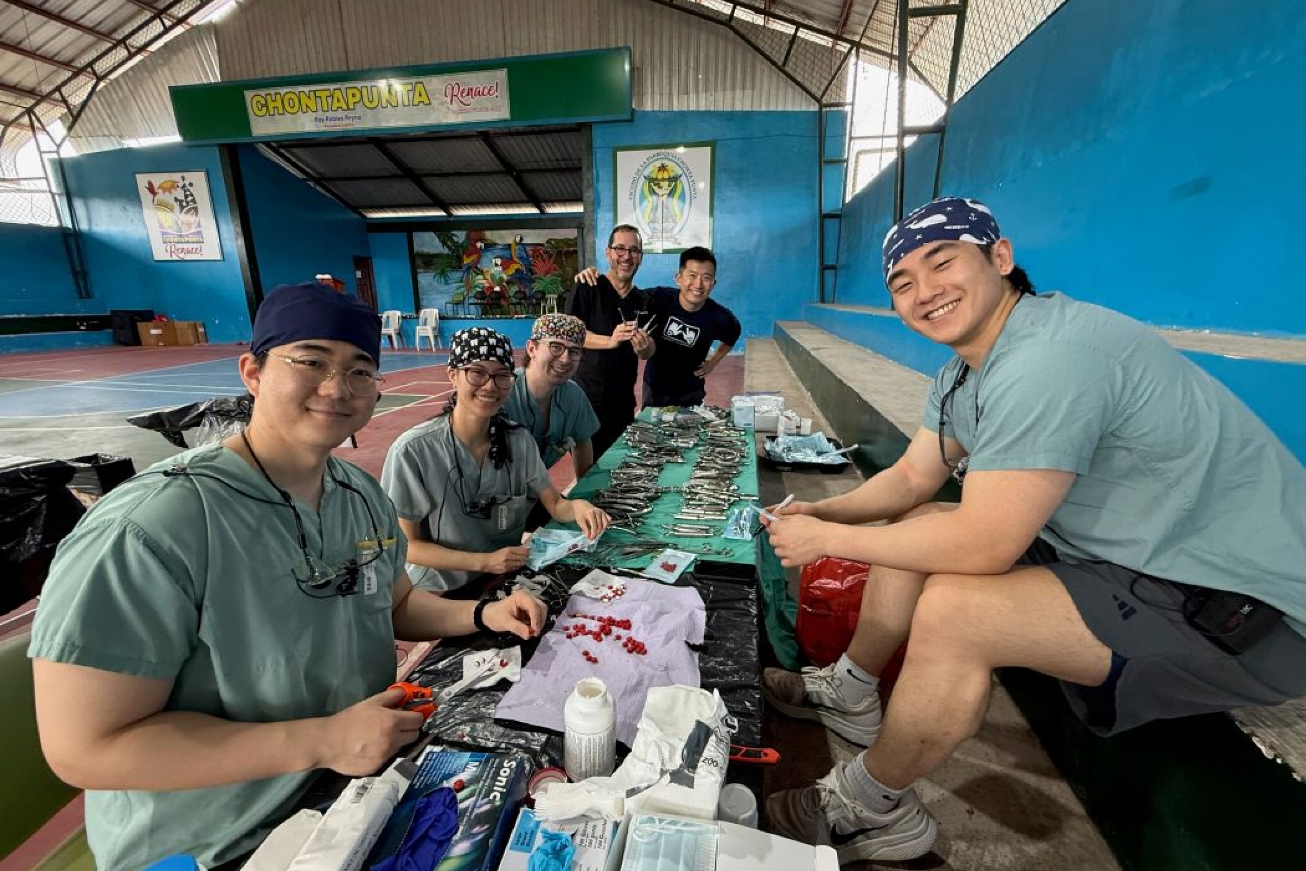 Six people pose for a photo in a gym. Dental instruments are on tables in front of them.