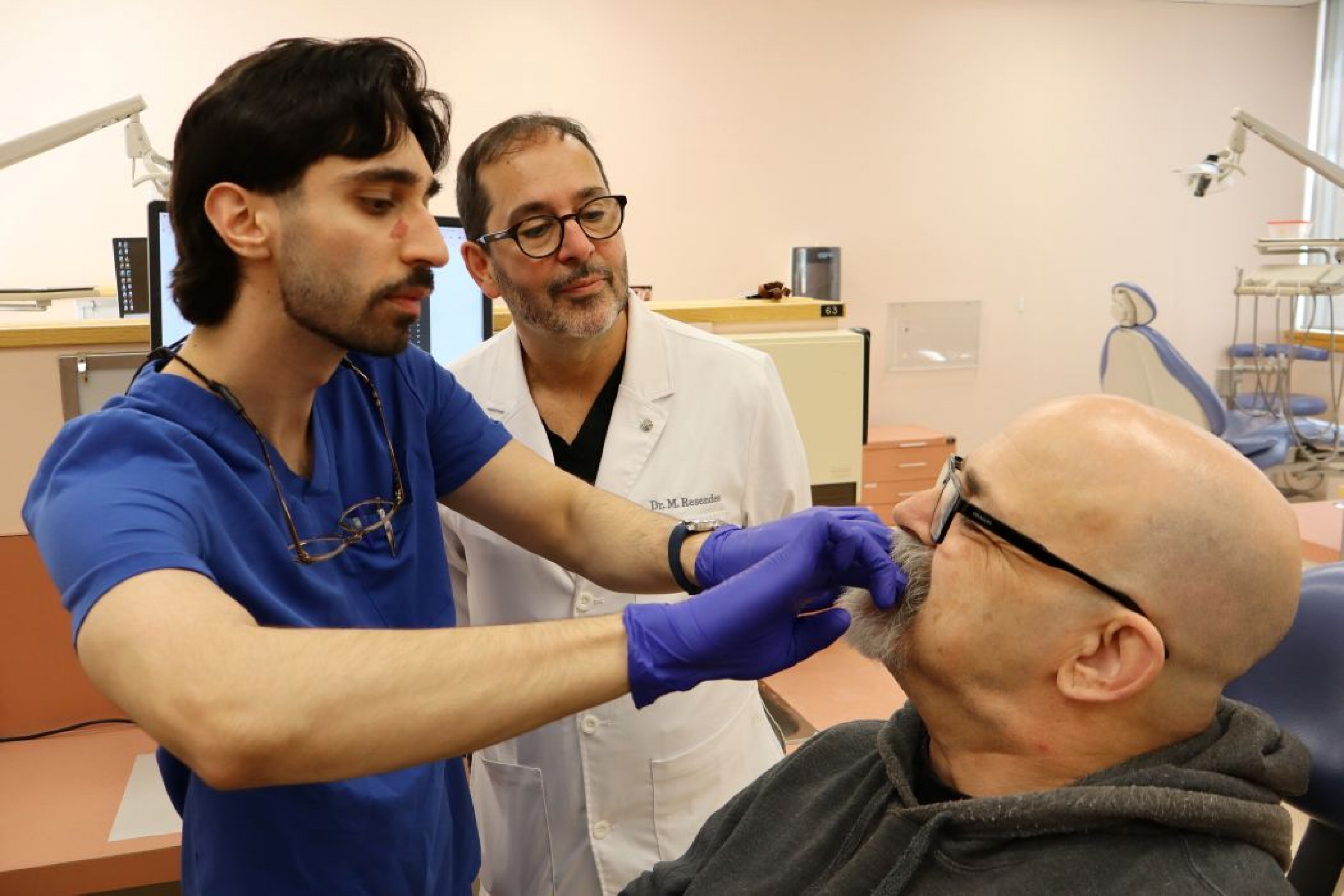 A dental student looks into a patient's mouth as an instructor watches.