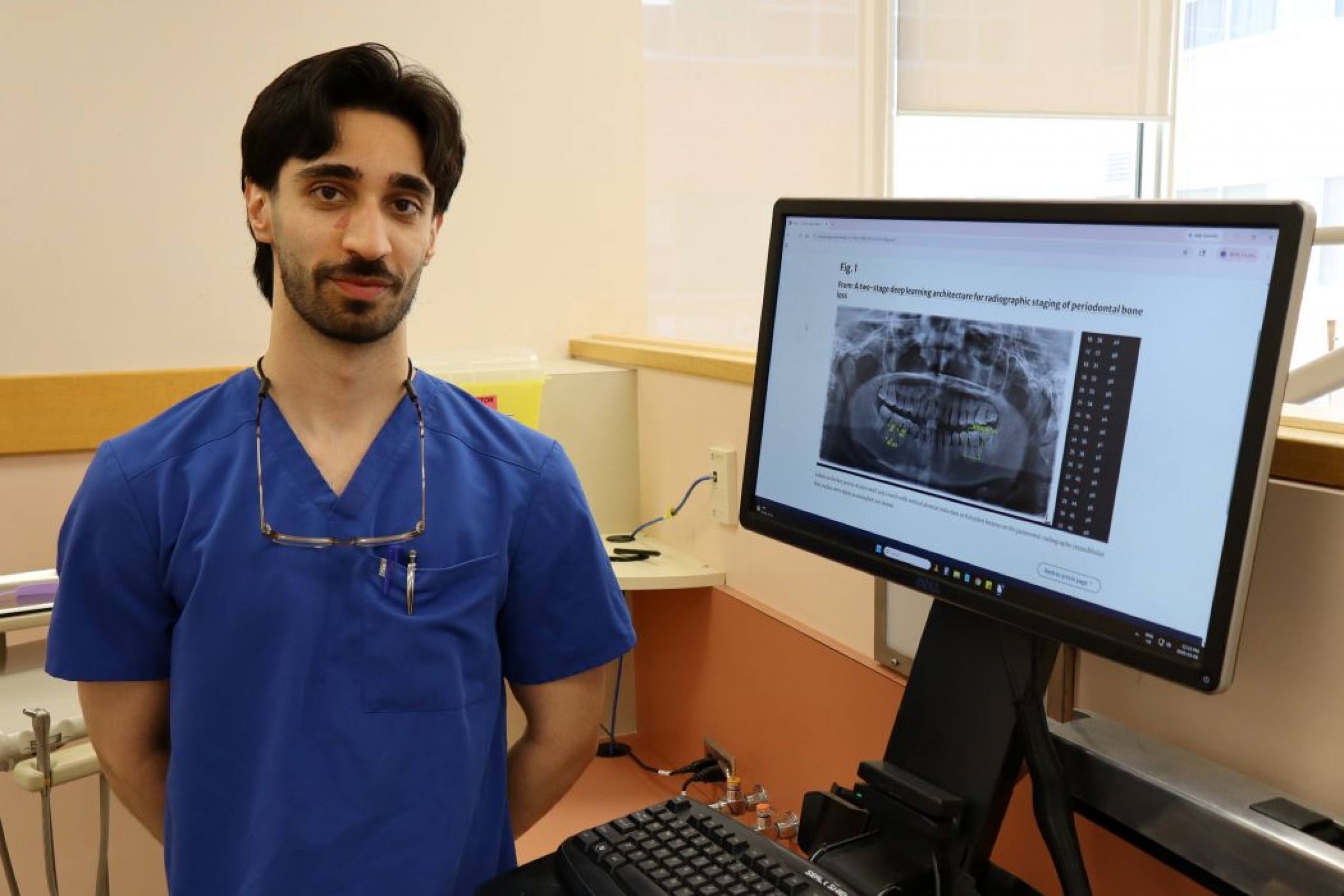 In a dental clinic, Hashim Abidullah stands next to a computer screen with an X-ray of a person's mouth.